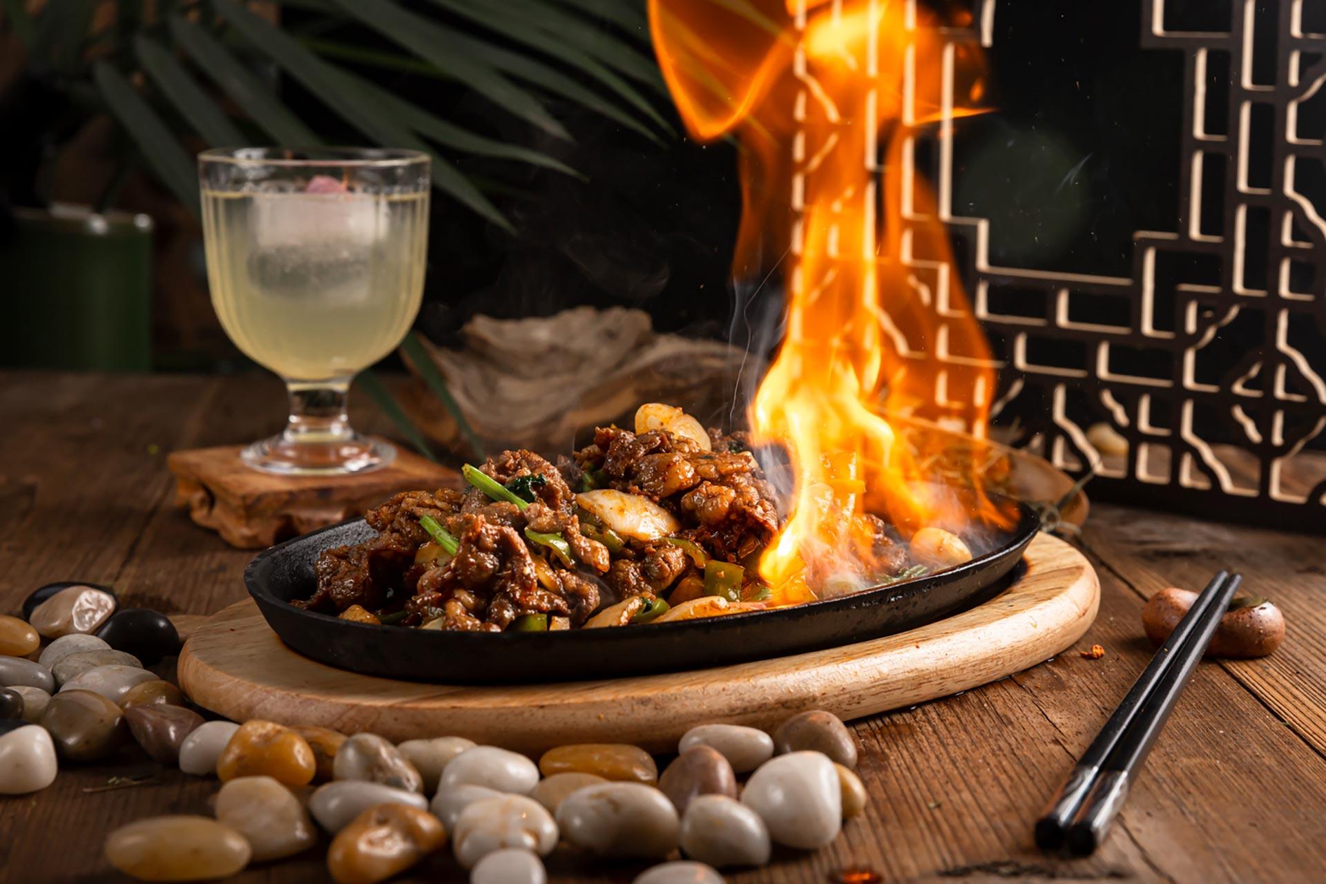 A sizzling platter of stir-fried meat and vegetables is set on a wooden table, with flames rising above the dish. A glass of a light-colored beverage and chopsticks are nearby, and decorative stones surround the plate.