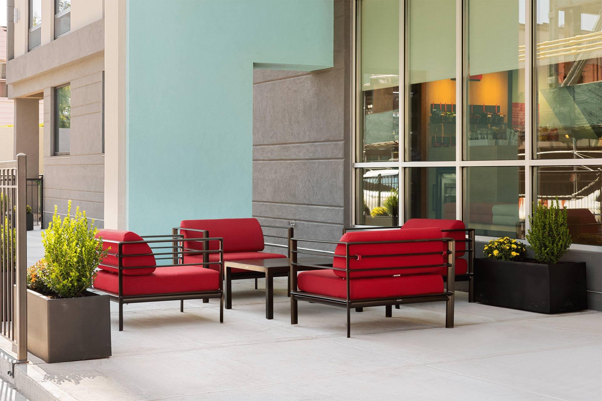 Modern outdoor patio with four red cushioned chairs around a wooden table, next to large windows. Two black planters with green and yellow plants decorate the space. The building exterior is light blue and gray.