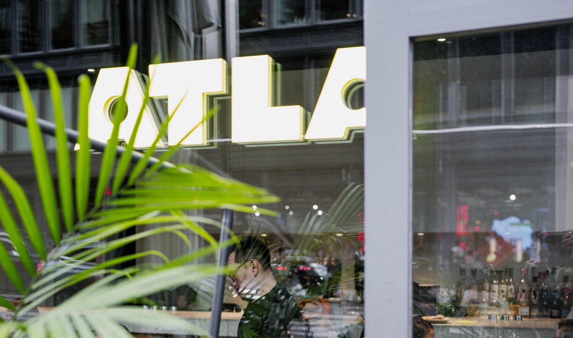 A man wearing glasses sits inside a modern café, partially obscured by a green plant. Large, bright letters spelling "OTLA" are visible on the glass window, with city reflections outside.