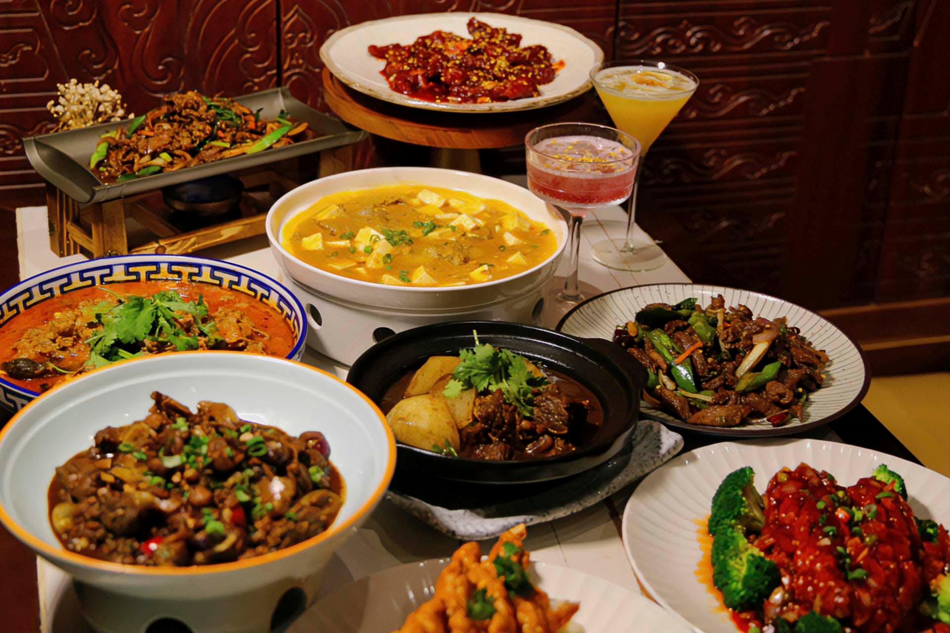 A table filled with various Chinese dishes, including a large tofu soup, stir-fried meats with vegetables, and vibrant garnishes. Two cocktails are placed alongside, and intricate wooden carvings are visible in the background.