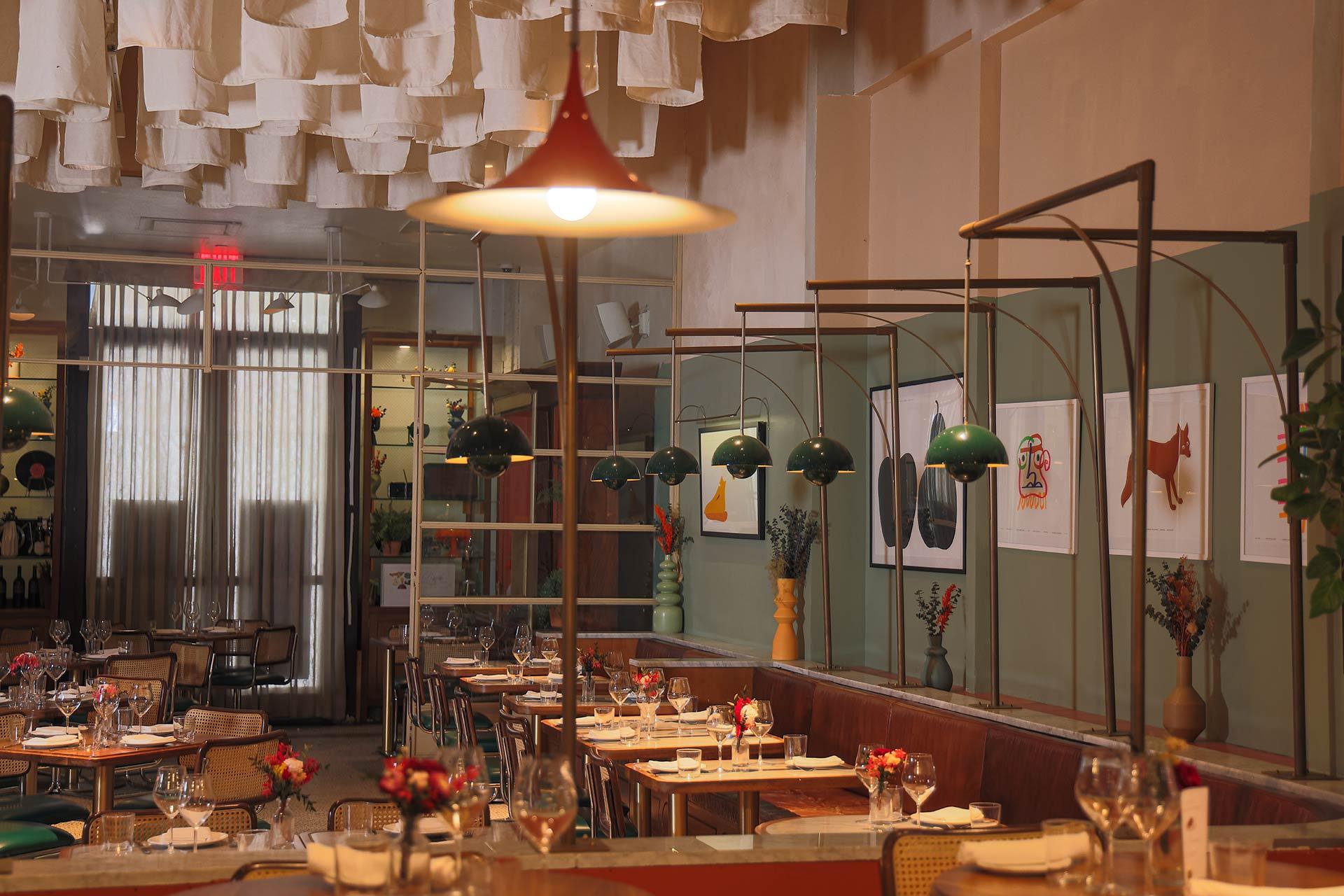 A stylish restaurant interior with set tables, green hanging lamps, wall art, and soft lighting. The ceiling features hanging fabric, and there are plants and flowers adding decorative touches throughout the space.