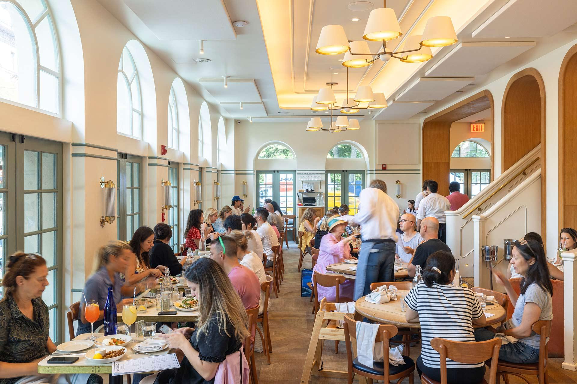 A busy, bright restaurant filled with people dining at wooden tables. Large arched windows let in natural light. A waiter serves food, and diners are engaged in conversation, creating a lively atmosphere.