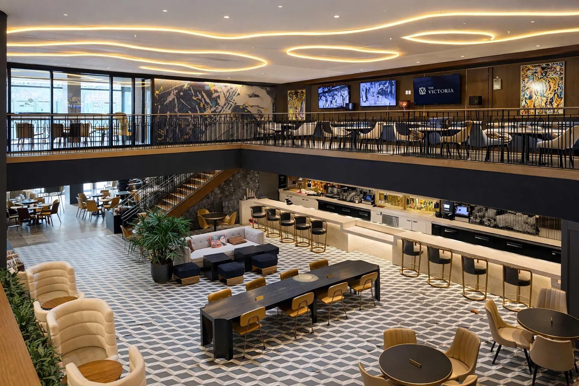 Modern, spacious lounge and bar area with geometric tile flooring, contemporary furniture, a long communal table, a staircase to a mezzanine level, and warm, wavy ceiling lights creating a welcoming atmosphere.