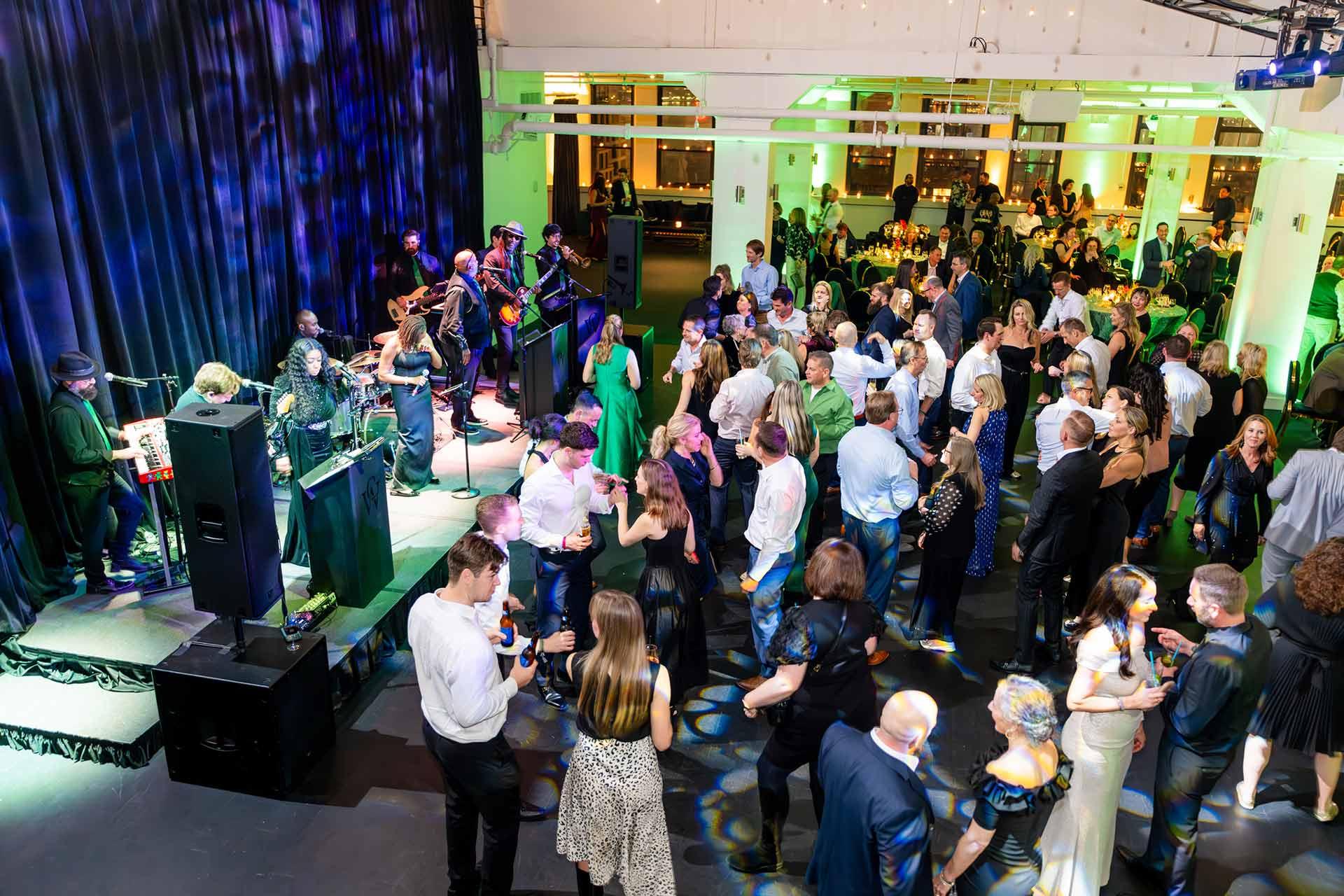 A lively indoor event with a live band performing on stage and a large crowd of people dancing, mingling, and socializing under colorful lights in a spacious venue.