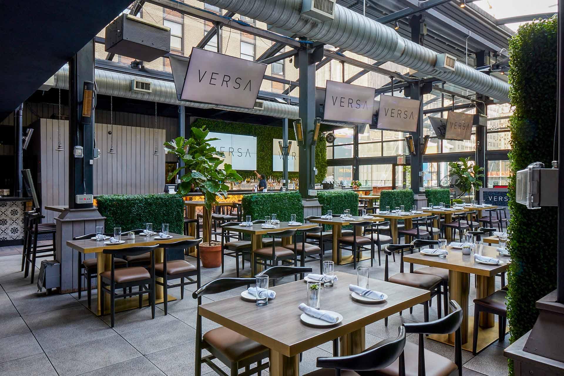 Modern restaurant interior with wooden tables, black chairs, and green plants. Several tables are set with glassware and napkins. Large VERSA signs are visible, and the space has an open, industrial feel with exposed ductwork.
