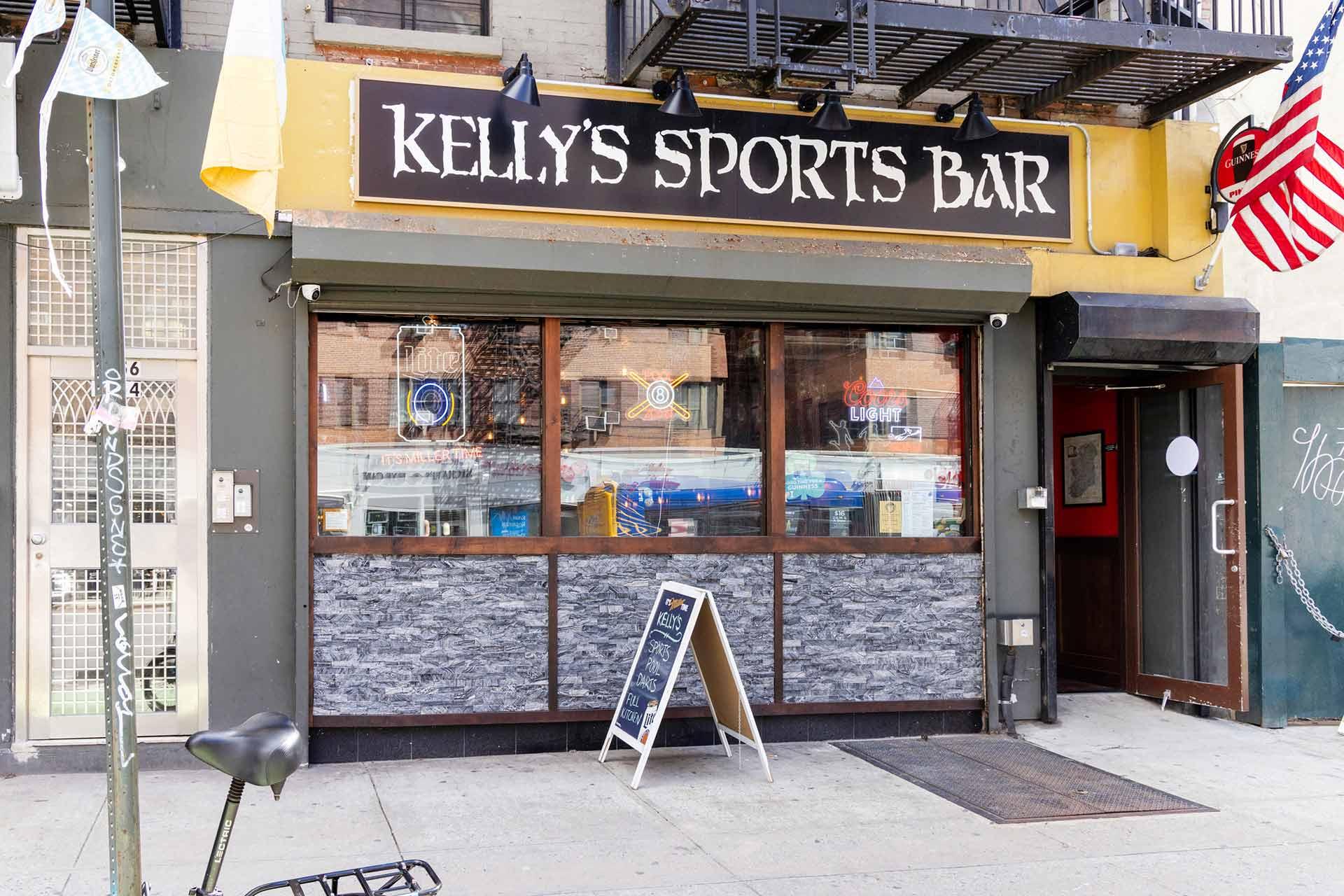 Exterior of Kelly’s Sports Bar with large front windows, a black and white sign above the entrance, a sandwich board on the sidewalk, and an American flag hanging by the door.
