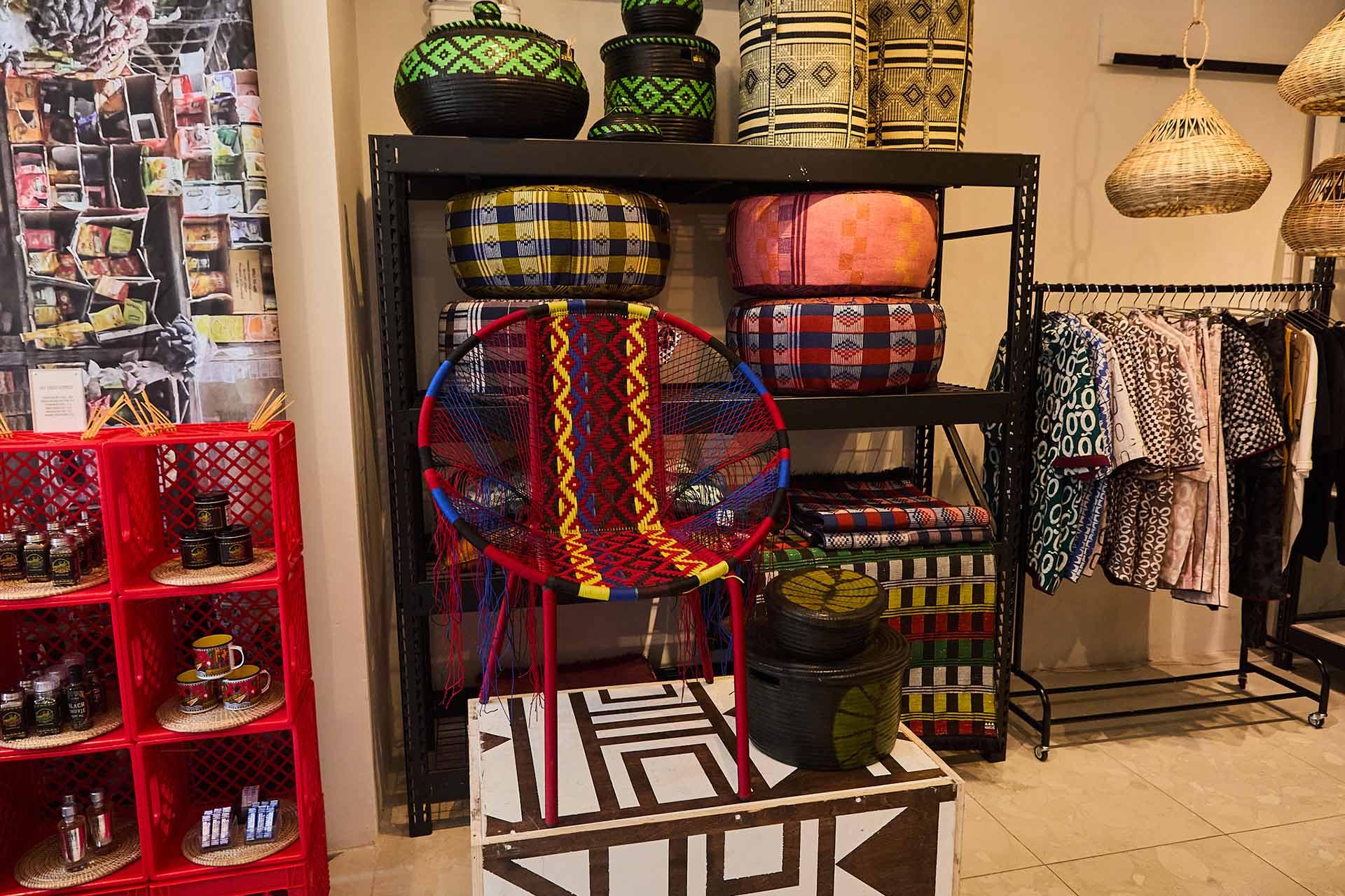 A colorful modern chair with a woven geometric design sits in front of shelves displaying patterned baskets, textiles, and pillows in a shop with vibrant décor and hanging woven lamps.