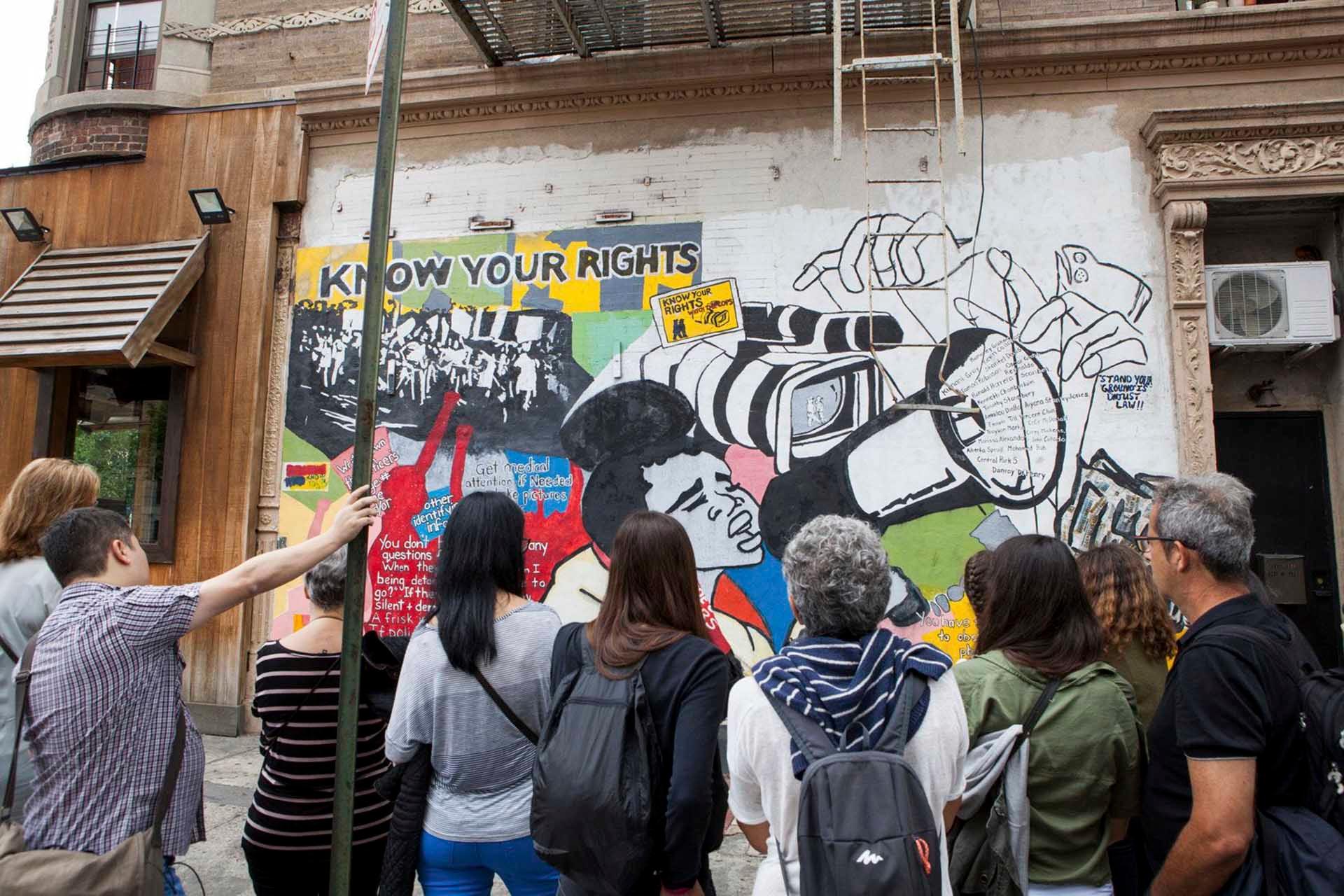 A group of people stand in front of a vibrant mural on a city building. The artwork includes text saying "KNOW YOUR RIGHTS" and depicts diverse figures and scenes, with bold colors and activist themes.
