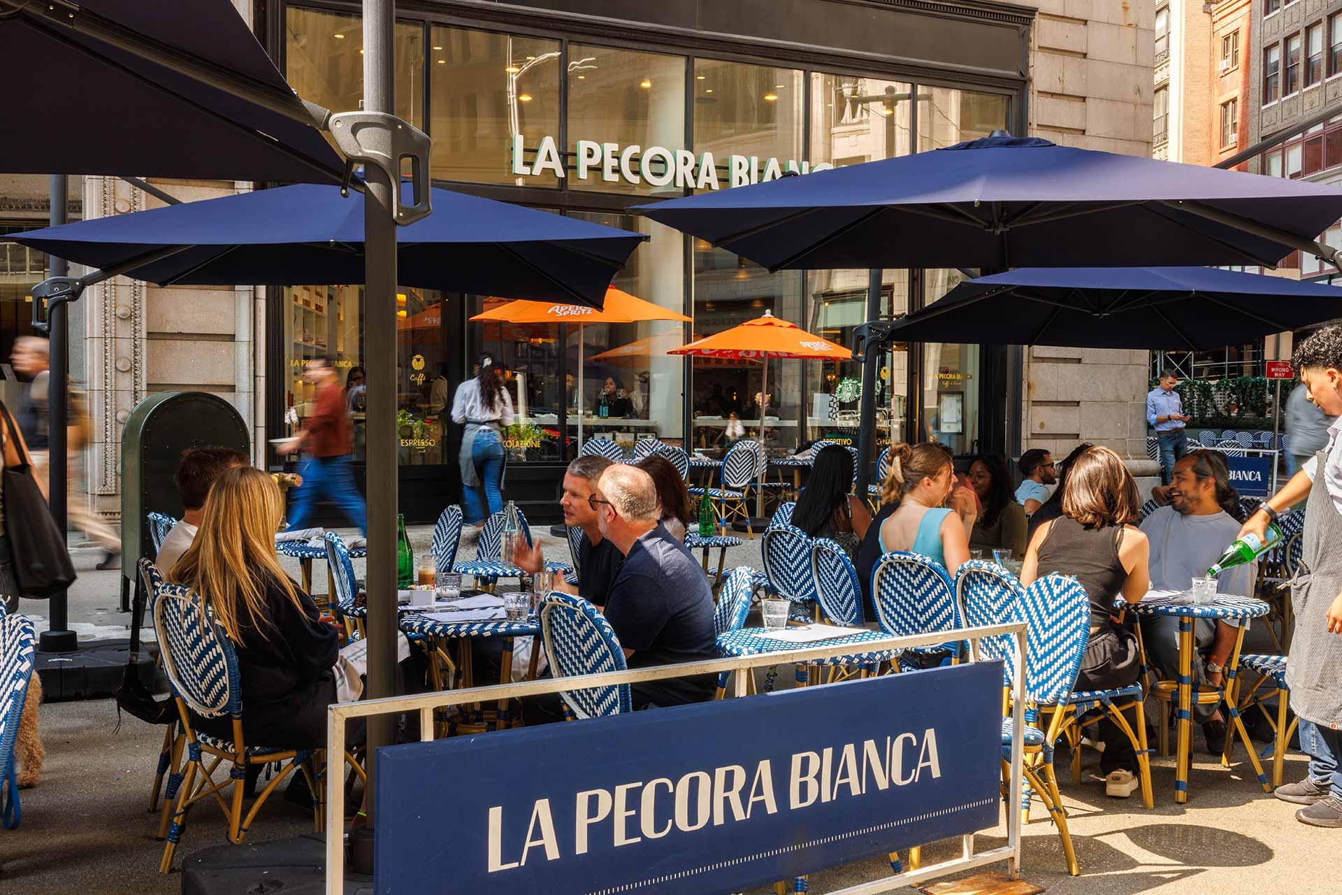 People dining at outdoor tables with blue umbrellas at La Pecora Bianca restaurant on a sunny day. The tables have blue and white checkered tablecloths, and the restaurant's sign is visible in the background.