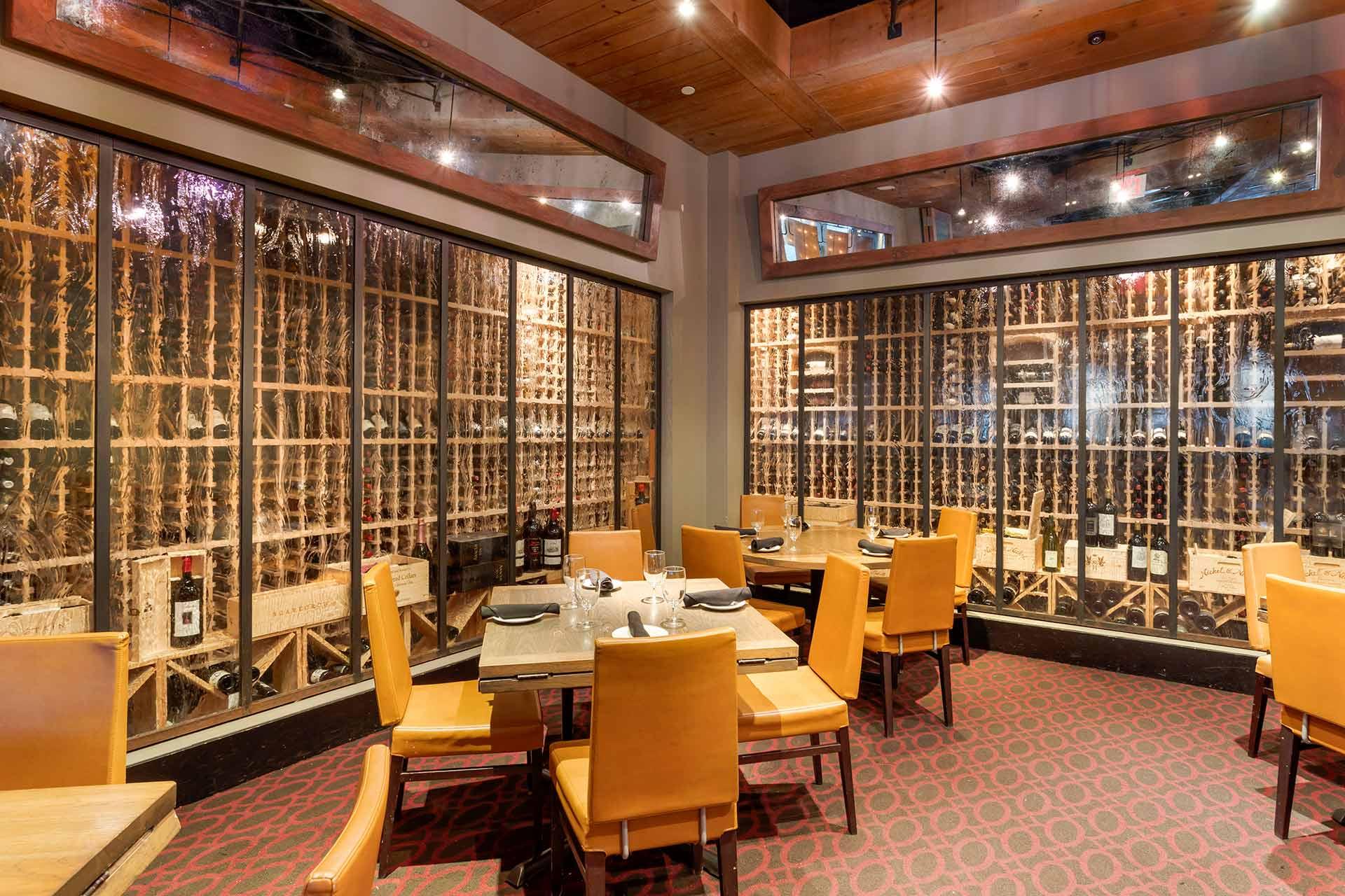 Private dining room with tan leather chairs around tables, place settings ready, and large glass windows displaying an extensive wine collection in wooden racks along two walls. Warm lighting and red-patterned carpet.