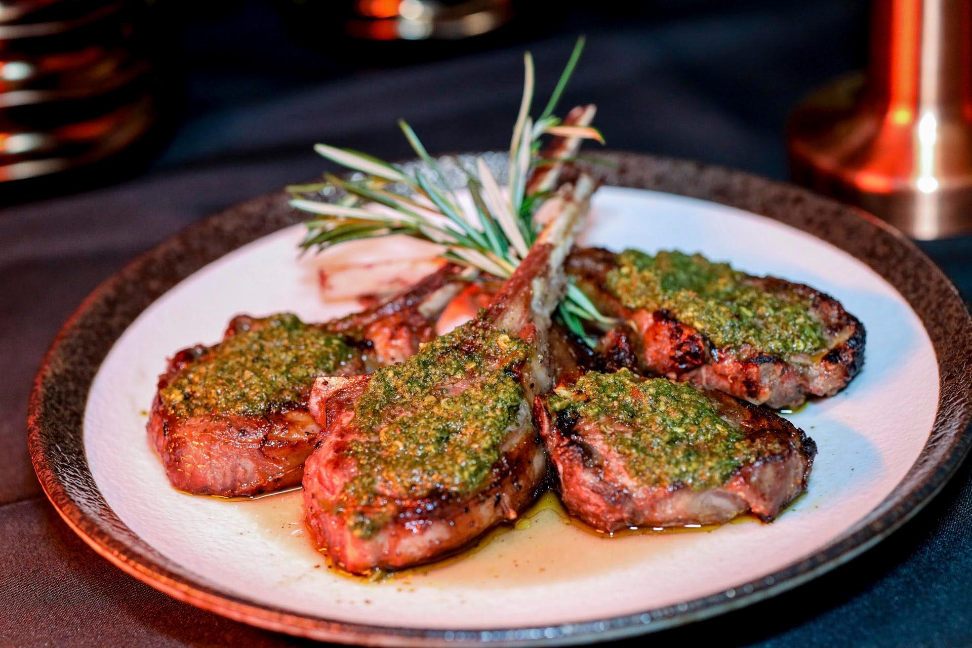 A plate of grilled lamb chops topped with a vibrant green herb sauce, garnished with sprigs of rosemary. The chops are arranged on a round, white plate with a dark rim, set against a dark background.