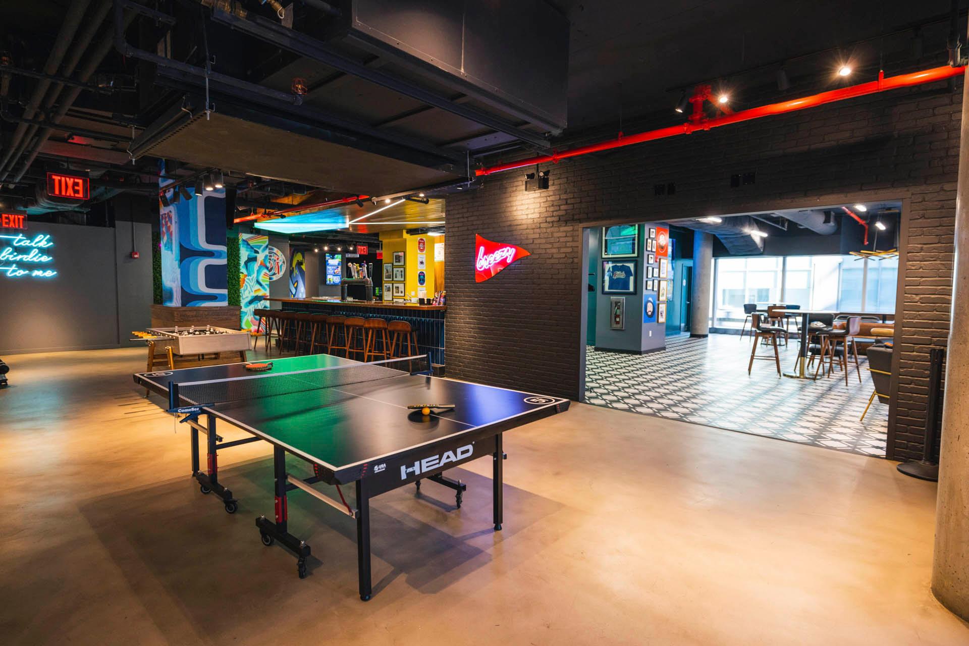 A modern recreational room features a ping pong table in the foreground, colorful wall art, a bar area with stools, and an open doorway leading to a bright dining space with tables and chairs.