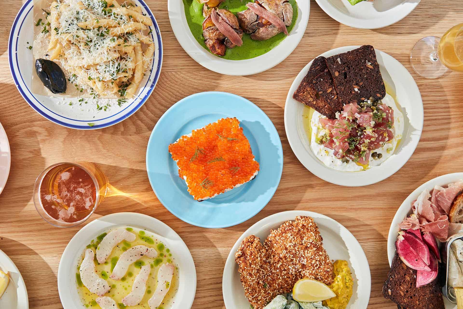 An overhead view of a wooden table filled with assorted dishes, including pasta, bread, seafood, raw fish, cured meats, a square of bright orange roe, and drinks. The table features a vibrant, colorful spread.