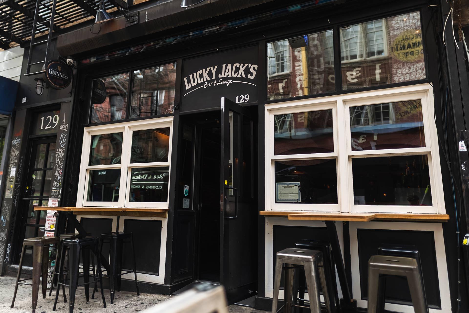 Street view of Lucky Jack’s Bar & Lounge with large windows, bar stools, a black exterior, and address number 129 displayed above the entrance. Reflections of nearby buildings are visible in the windows.