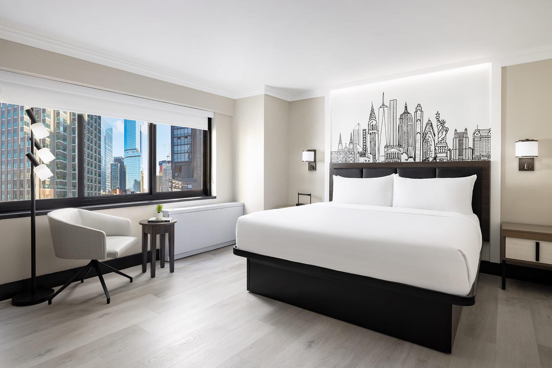 A modern hotel room with a large bed, minimalist furniture, a desk and chair by the window, and a city skyline wall mural above the bed. Large windows offer views of city buildings outside.