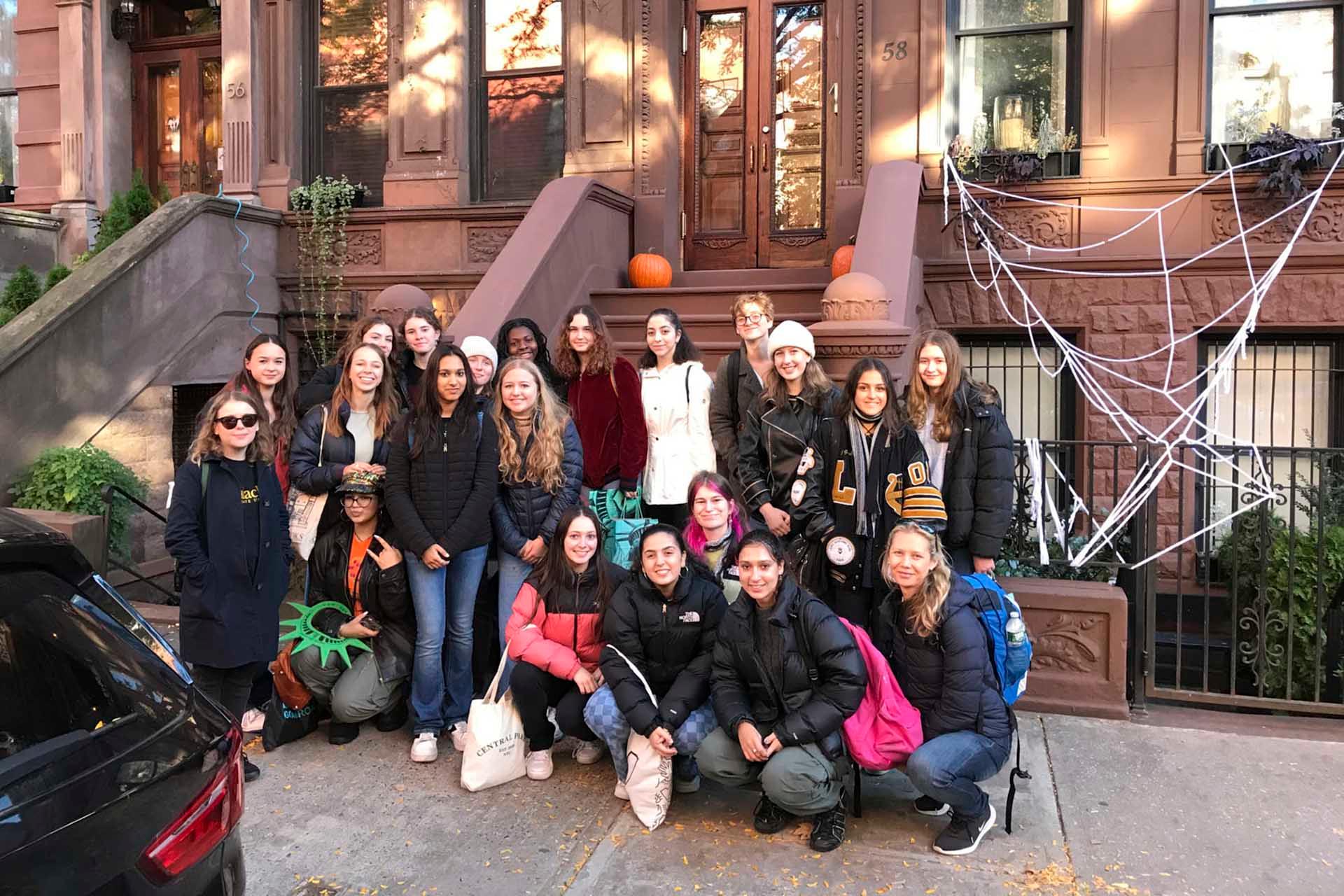 A group of twenty people, mostly women, pose together on a sidewalk in front of a brownstone building. The building is decorated with pumpkins and a spider web for autumn or Halloween. Some individuals are wearing jackets and holding bags.