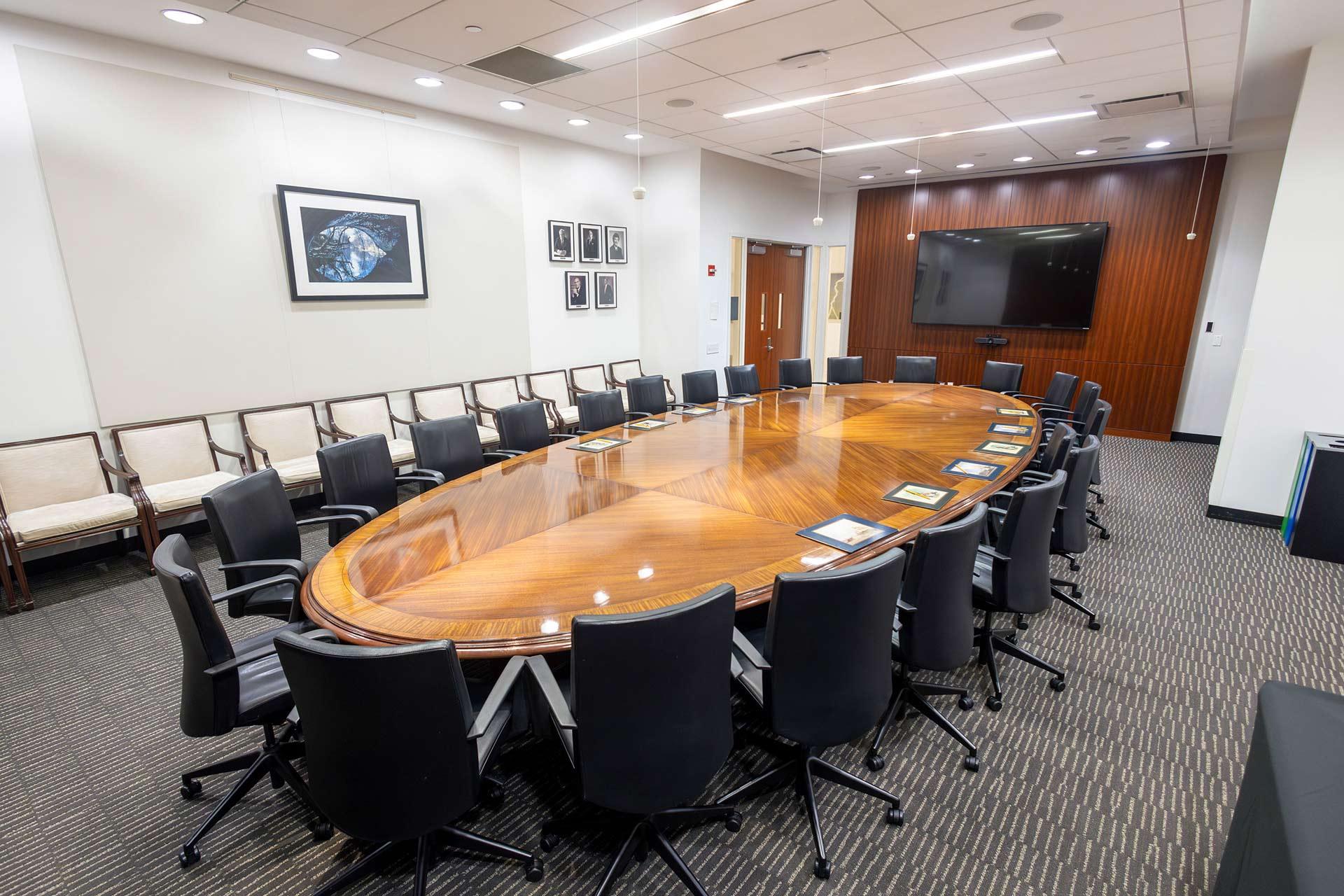 A modern conference room with a large oval wooden table surrounded by black office chairs, a large wall-mounted screen, framed art on the walls, and beige chairs along one side.
