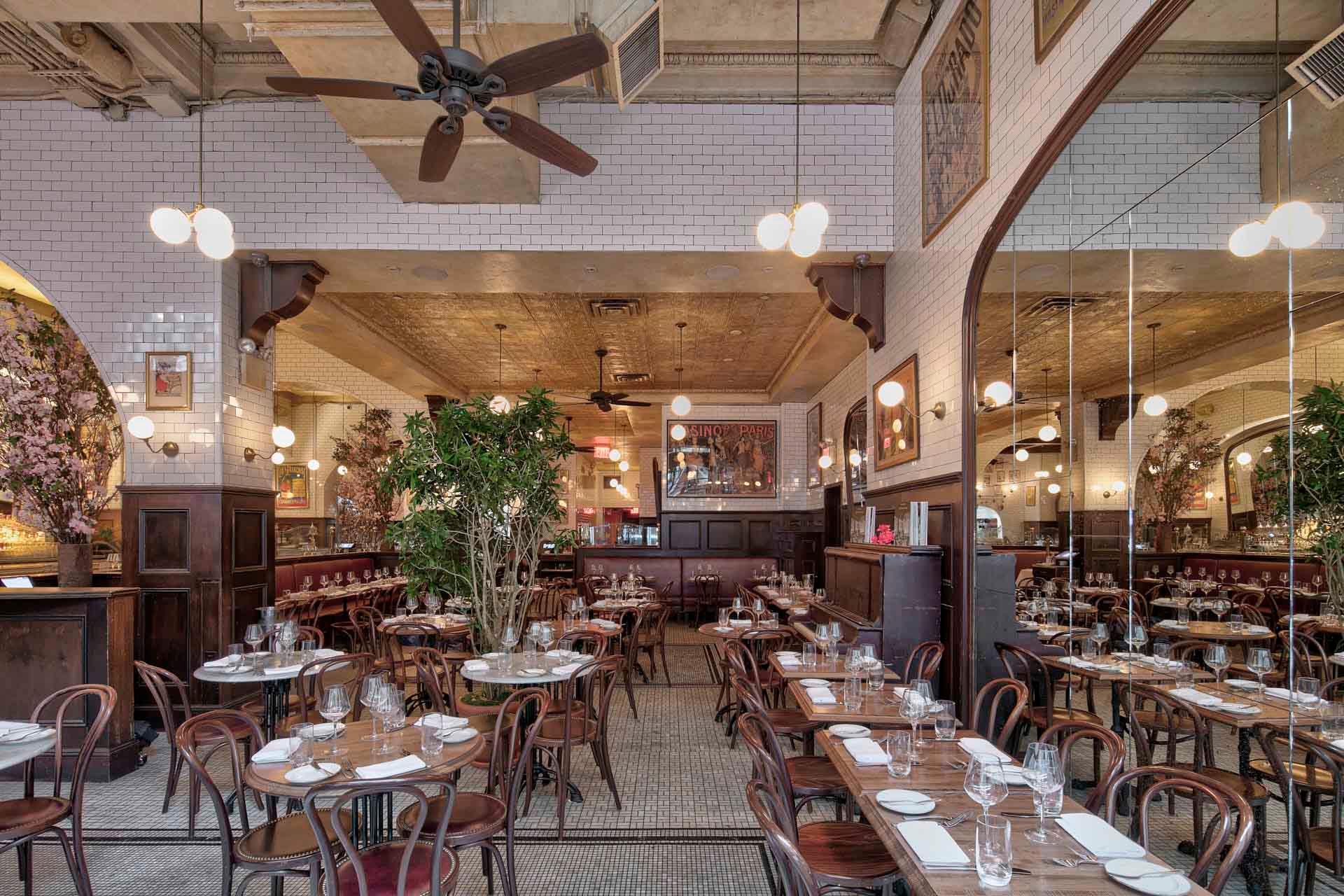 Spacious restaurant interior with high ceilings, white subway tile walls, and elegant lighting. Wooden tables and chairs are neatly arranged, each set with glassware and white napkins. Large potted plants add greenery to the warm atmosphere.