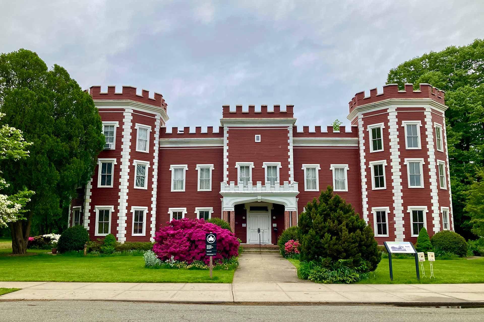 A large, red building with white trim and castle-like towers stands behind a sidewalk, surrounded by green trees, grass, and blooming pink bushes.