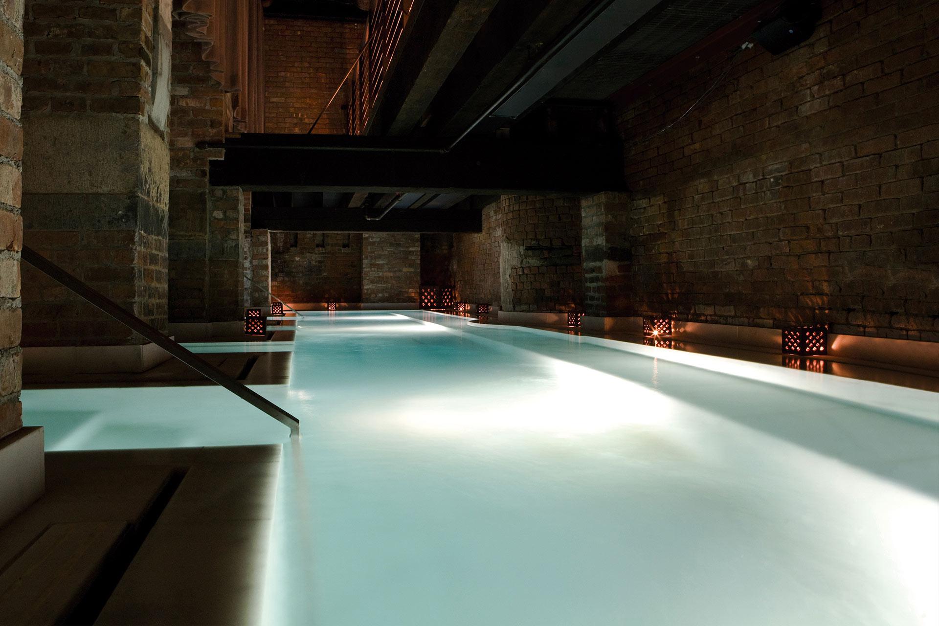 A long indoor pool with glowing white water, surrounded by exposed brick walls and subtle lantern lighting, creating a calm and atmospheric setting under a metal walkway.