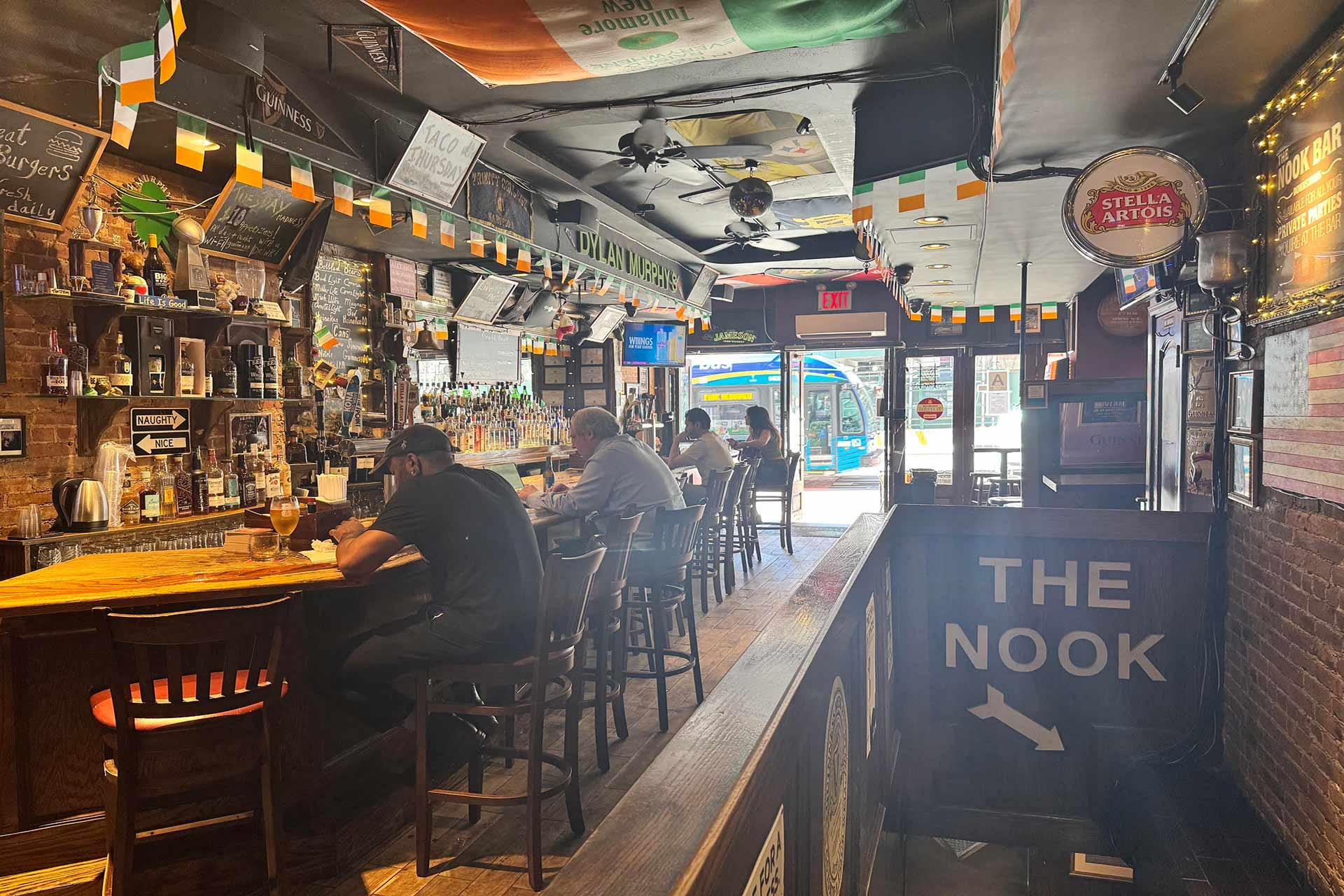 People sit at a wooden bar inside a dimly lit Irish pub decorated with flags and lights. Bottles line the shelves, and a sign reading “THE NOOK” hangs on the right. Sunlight streams through the front windows.