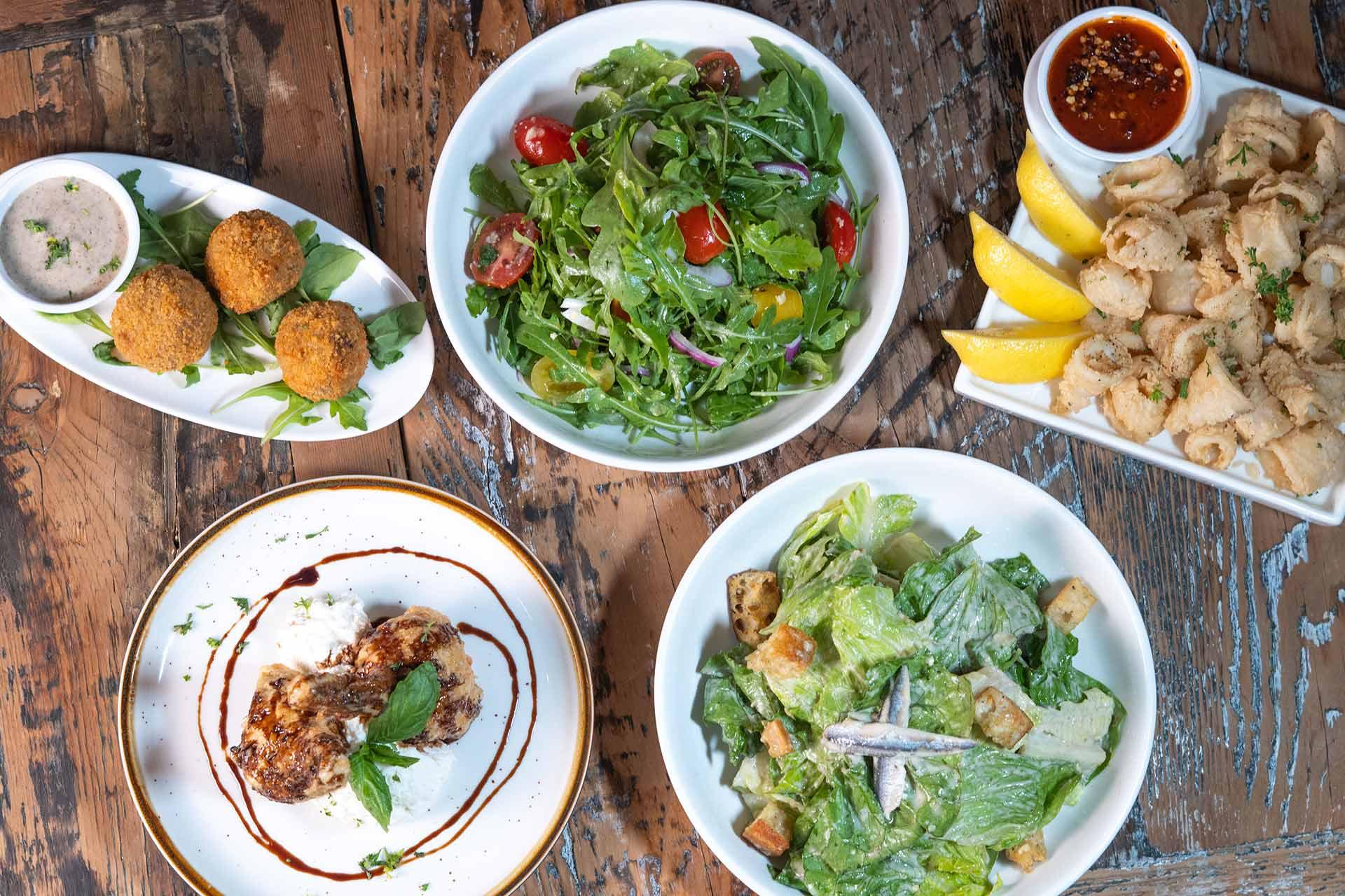 An overhead view of assorted dishes on a wooden table, including two fresh salads, a plate of fried appetizers with lemon wedges, falafel with dip, and a dish topped with fresh herbs and balsamic glaze.