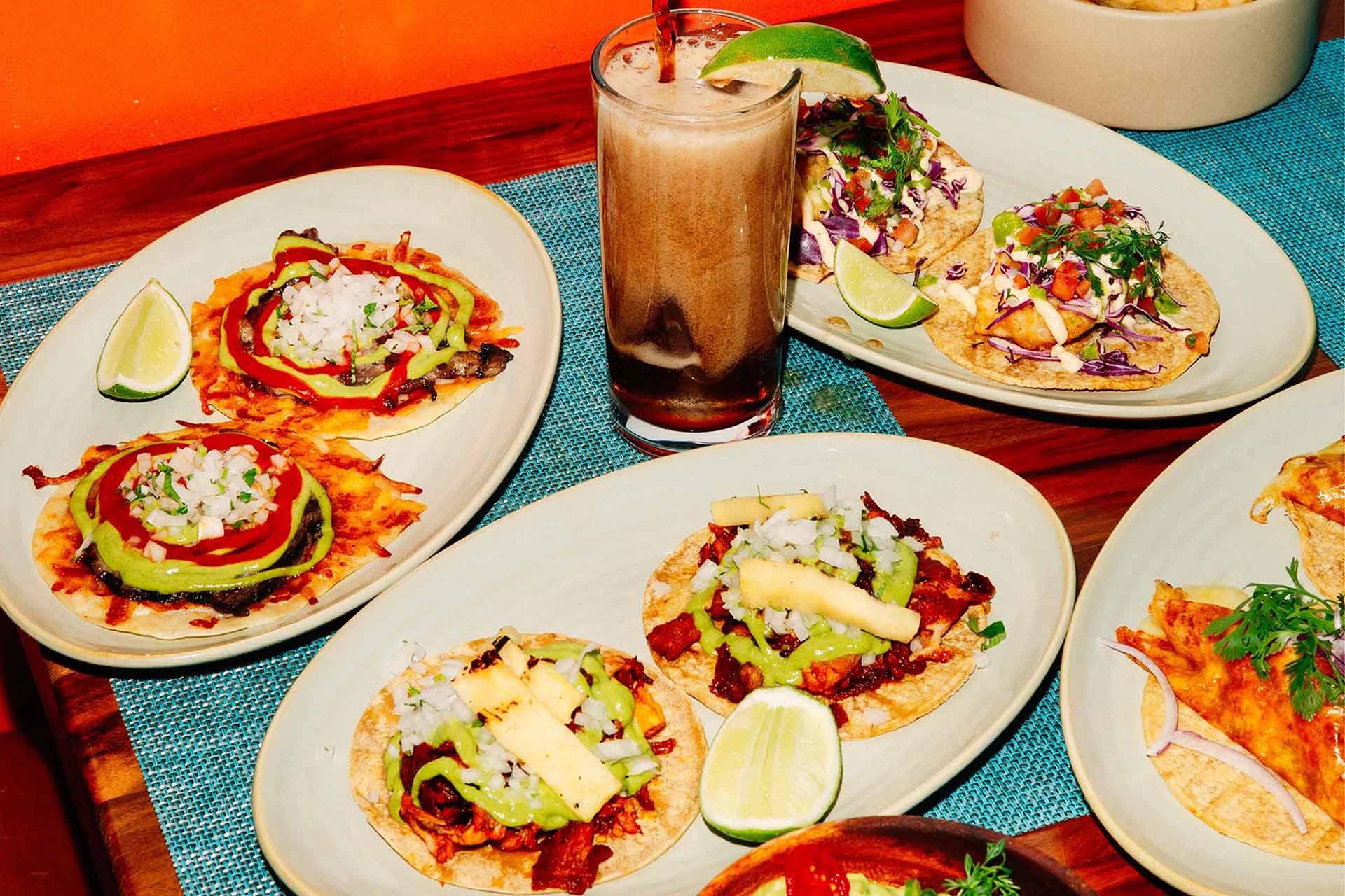 A table with various tacos topped with vegetables and pineapple, lime wedges on the side, and a tall glass of soda with a lime wedge garnish.