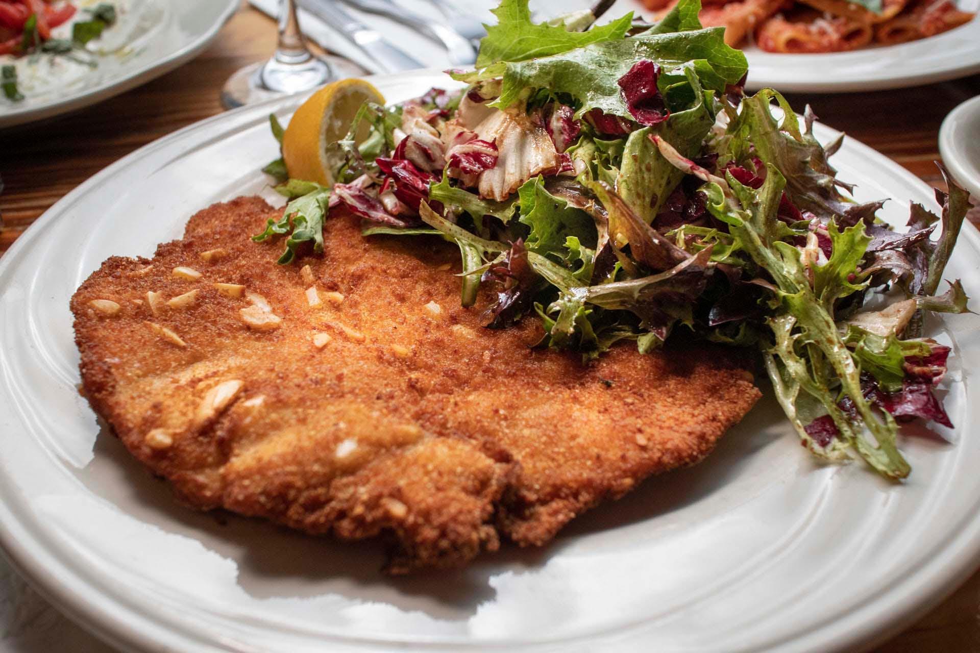 A crispy breaded schnitzel served on a white plate, topped with a fresh mixed green salad and accompanied by a lemon wedge. The dish is set on a wooden table, with parts of other plates visible in the background.