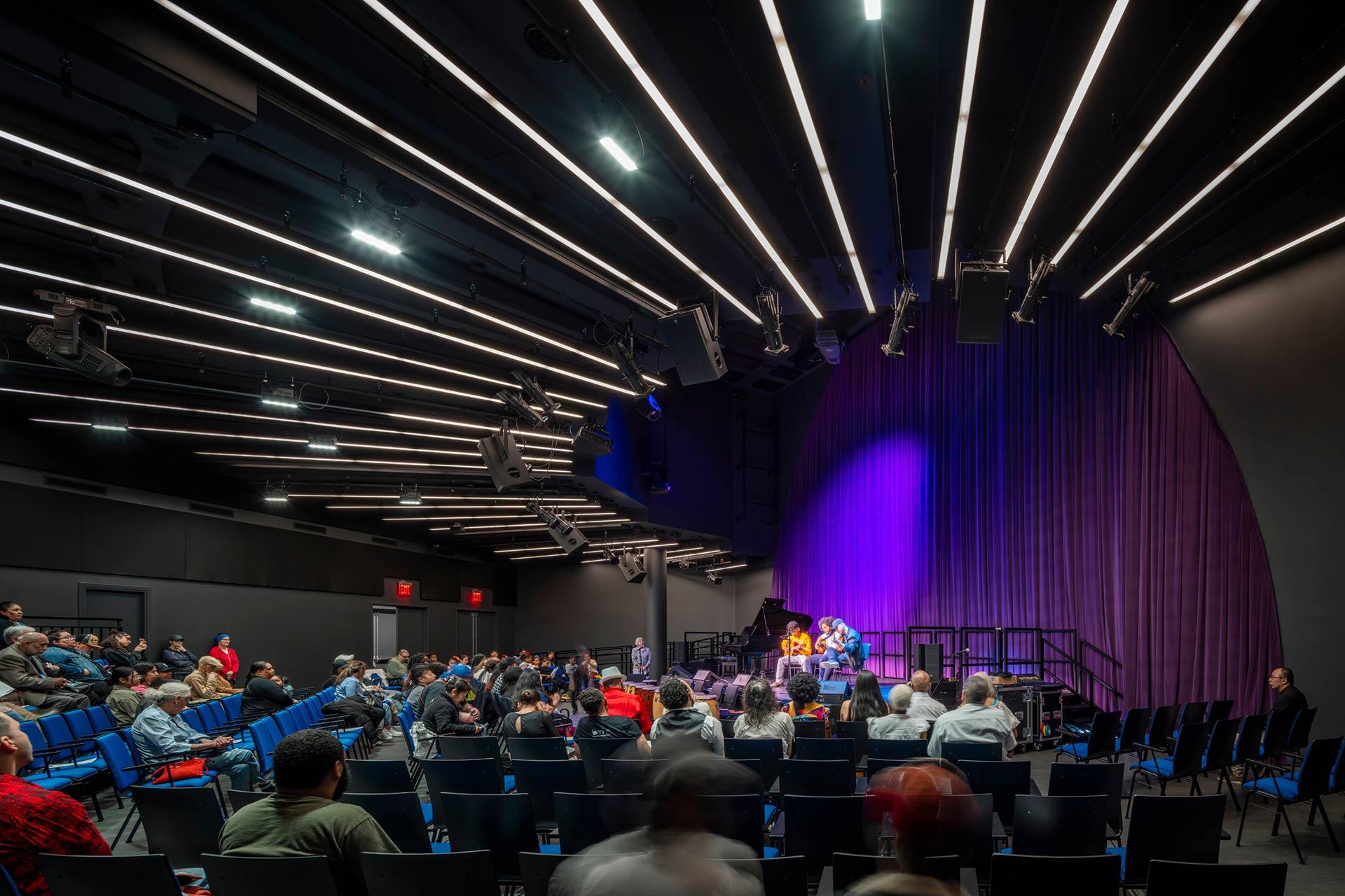 A modern auditorium with people seated, facing a stage lit with purple lighting. Several panelists sit on stage in front of a closed curtain. The ceiling features rows of linear lights. Some attendees are still arriving.