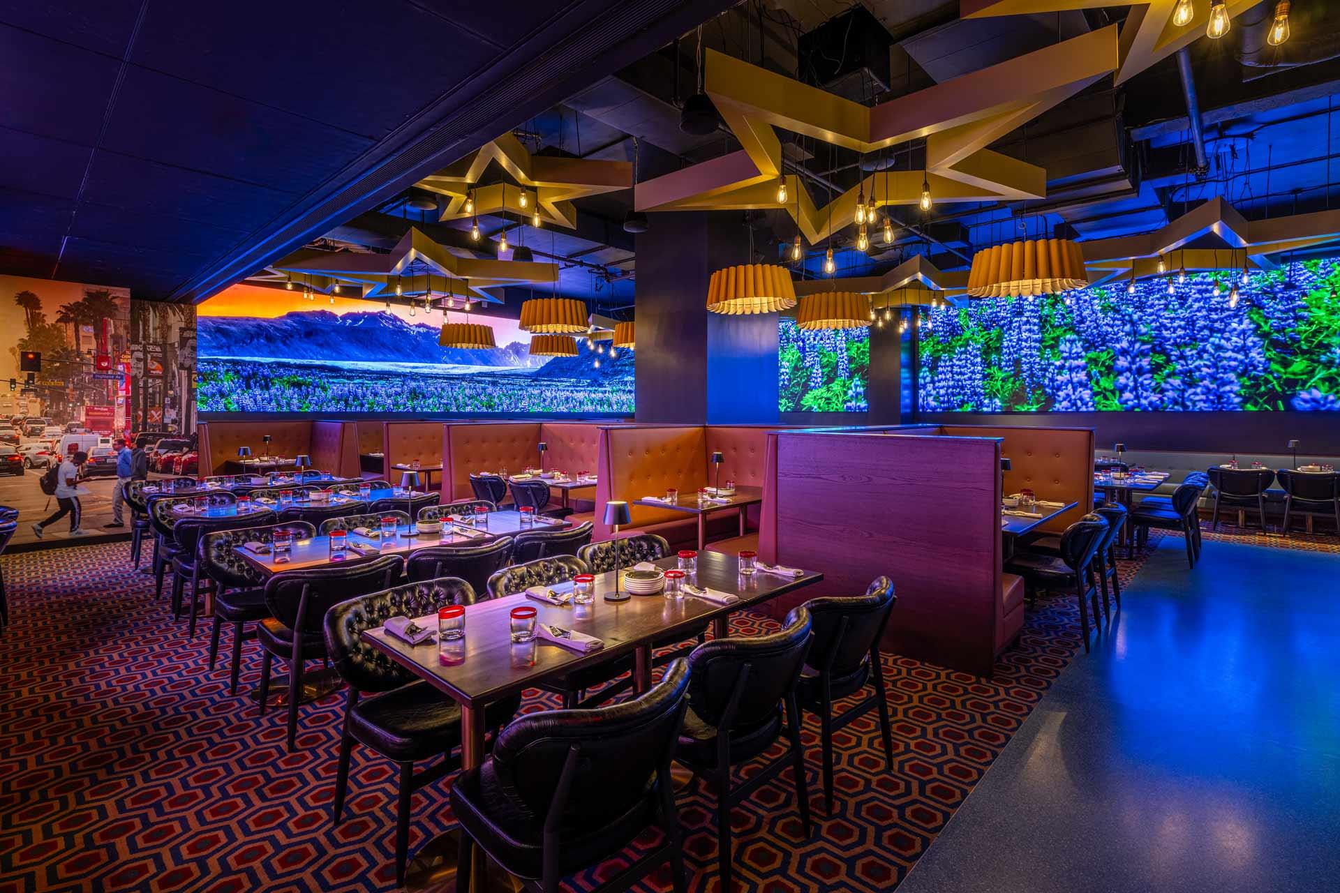 A vibrant restaurant interior featuring tables set with glasses and tableware. The decor includes geometric ceiling designs, warm lighting, and an illuminated wall with greenery. The overall ambiance is modern and lively.