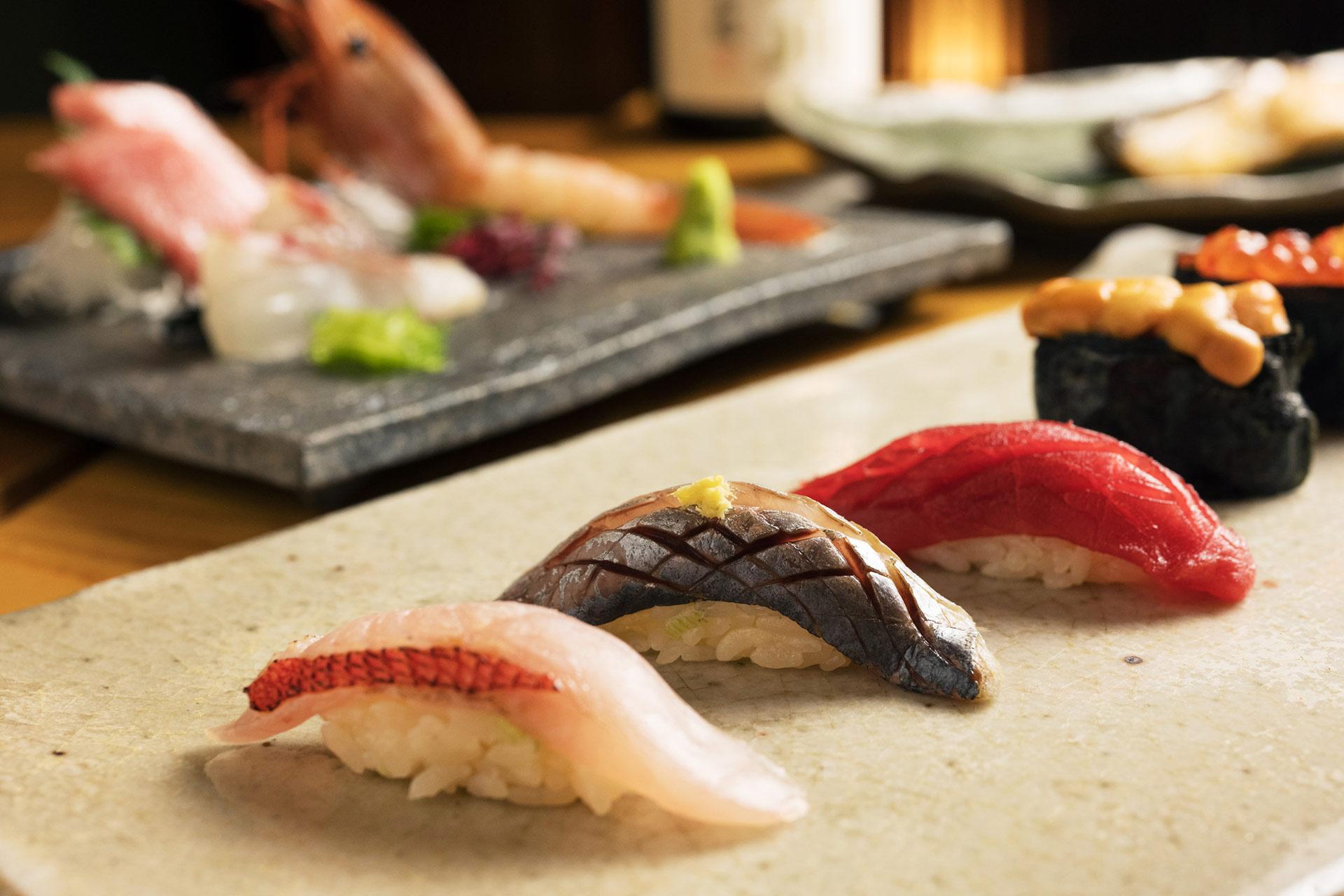 A close-up of assorted sushi pieces, including nigiri with different types of fish, arranged on a stone plate with more sushi and sashimi in the blurred background.