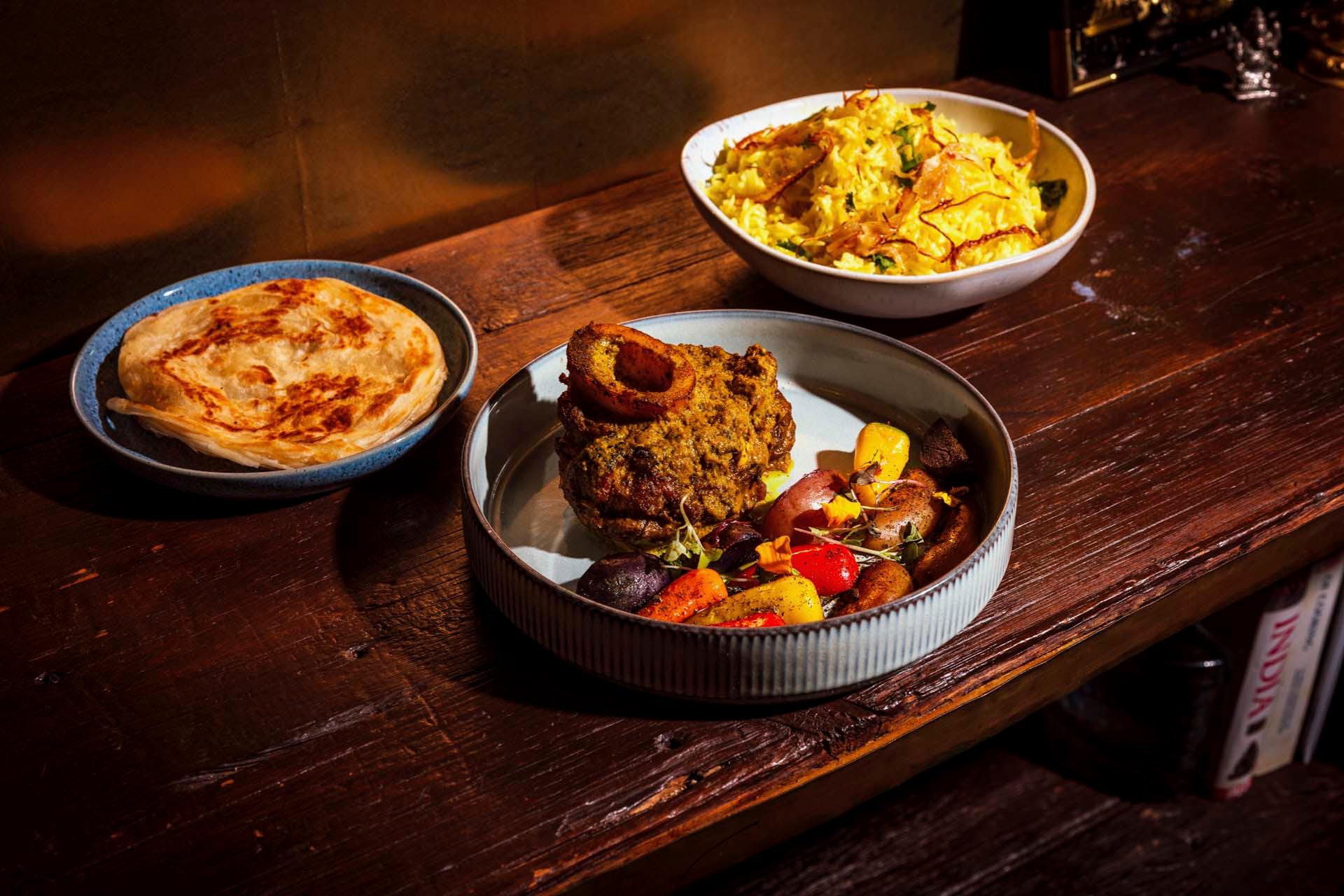 A wooden table holds three dishes: a plate with a crispy flatbread, a bowl with yellow rice garnished with herbs and crispy toppings, and a plate featuring a piece of roasted meat with assorted vegetables.