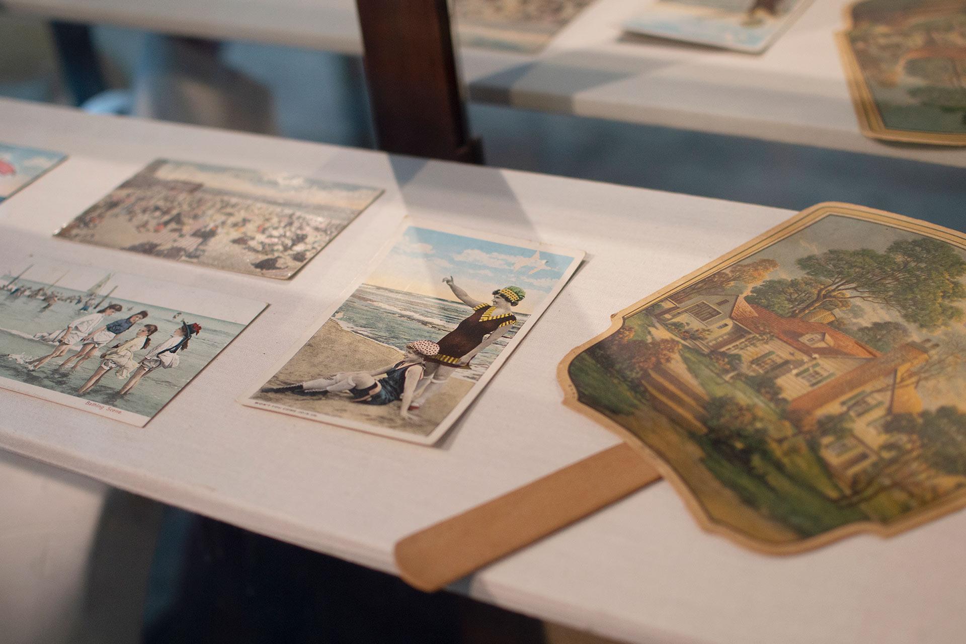 Several vintage postcards and a hand fan with an illustration of a house are displayed on a white surface. The postcards feature beach scenes and people near the ocean, evoking a nostalgic, summery feel.