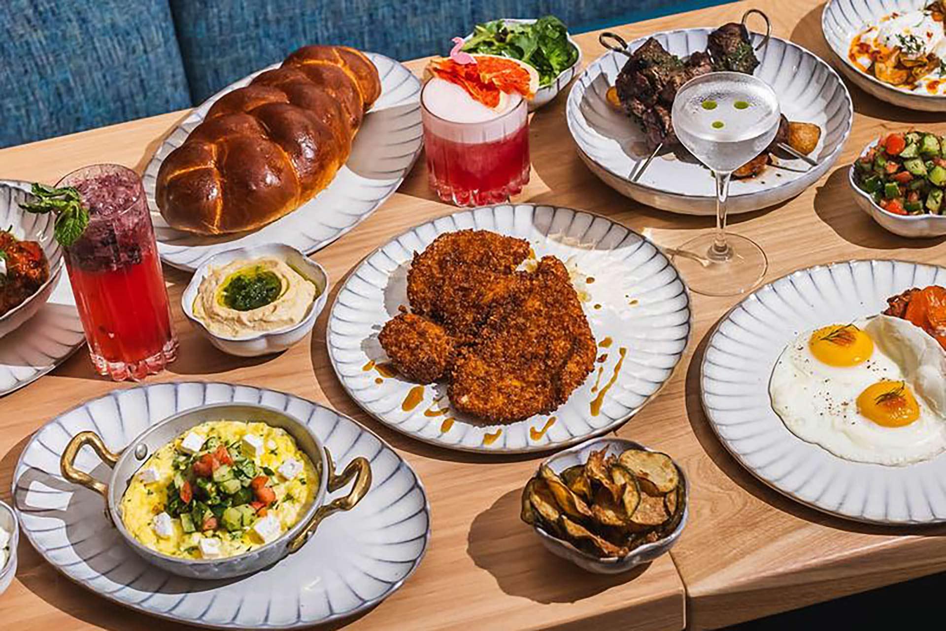 A table set with various dishes including fried chicken, sunny-side-up eggs, braided bread, grilled skewers, small salads, fried potato slices, and colorful cocktails, all served on white plates.
