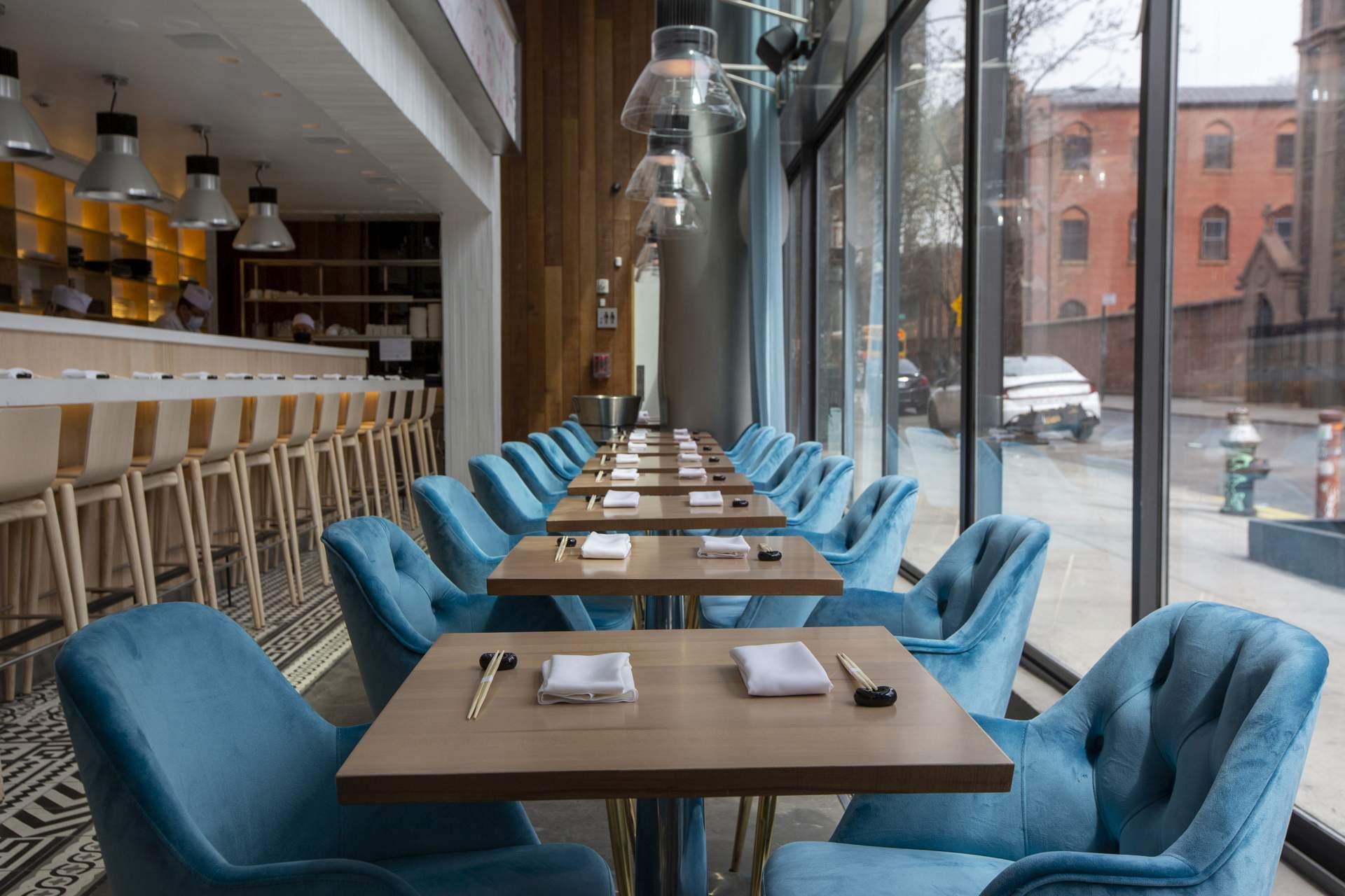 A modern restaurant interior with tables set for dining, featuring plush blue chairs, wooden tables with napkins and utensils, and large floor-to-ceiling windows overlooking a city street.