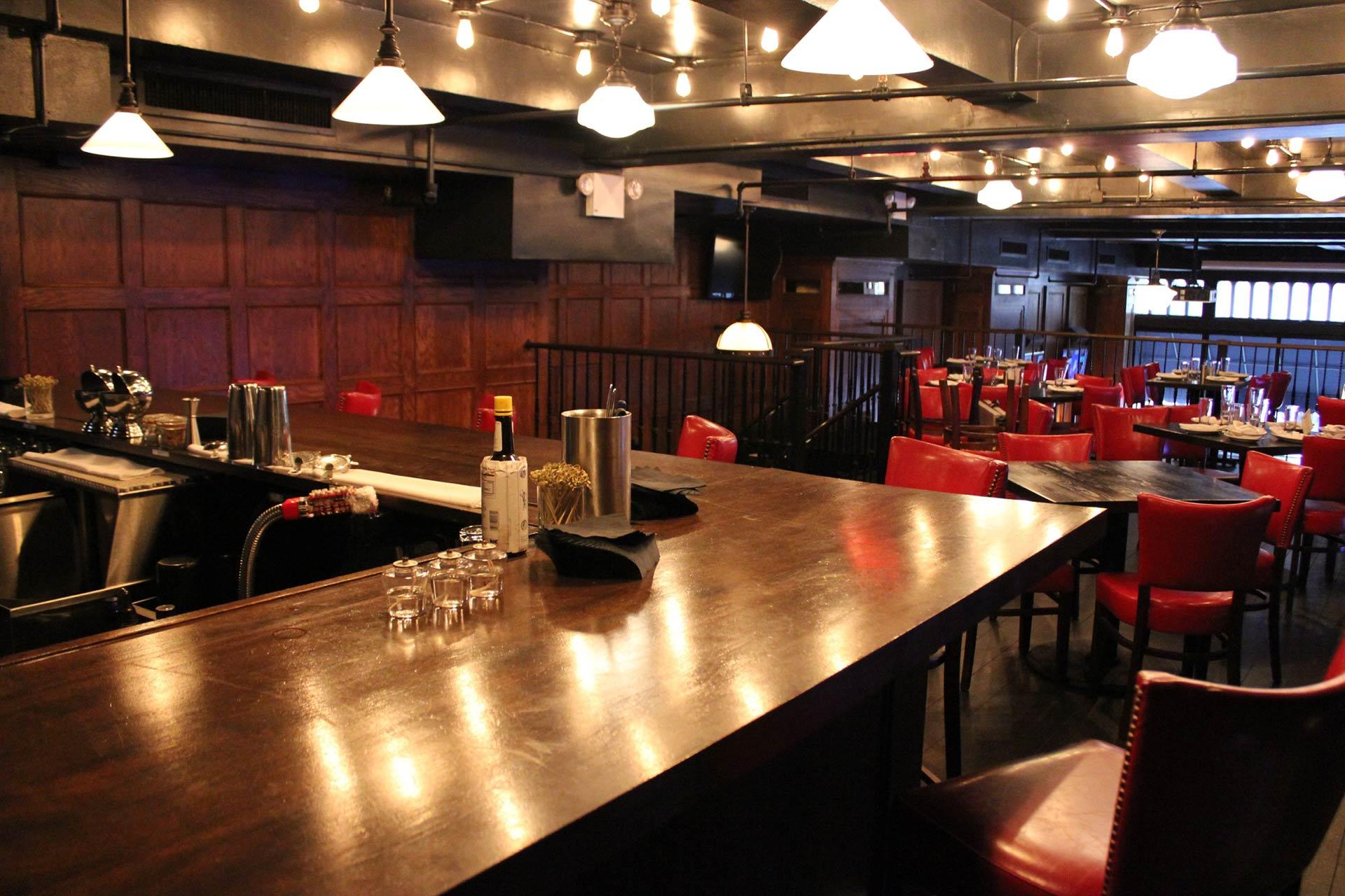 A dimly lit bar with wooden walls, a polished countertop, red chairs, and tables. Bottles and glasses are on the bar, and string lights hang from the ceiling, creating a cozy atmosphere.