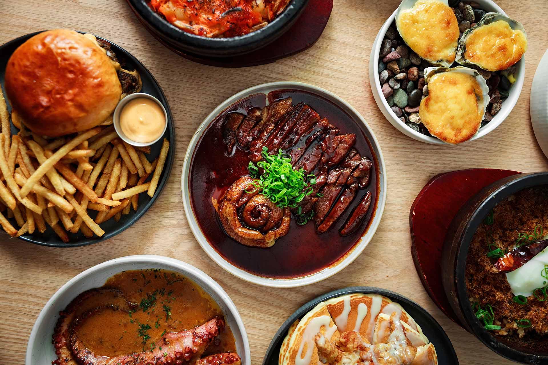 An overhead view of various dishes, including steak with sauce and green onions, a burger with fries, baked oysters, grilled octopus, and other assorted foods on a wooden table.