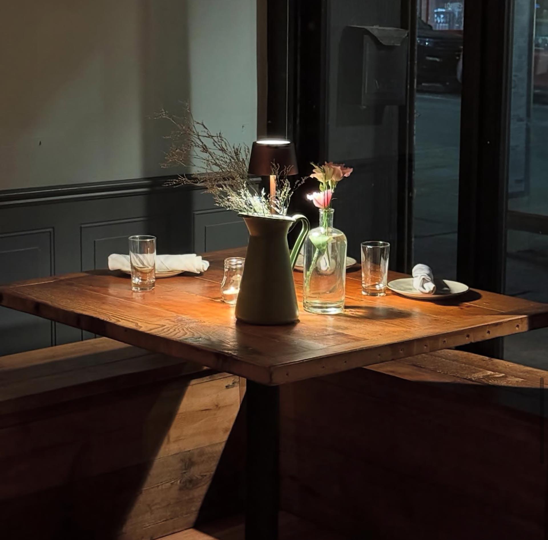 A cozy wooden table set for two with glasses, a green ceramic pitcher, a glass vase with flowers, and a small lamp, bathed in warm light near a window at night.