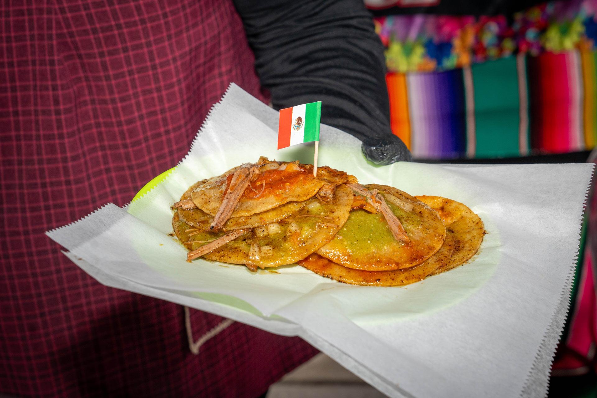 A person holds a paper plate with four small tortillas topped with shredded meat and green and red sauces, decorated with a small Mexican flag toothpick. The background includes colorful fabric.
