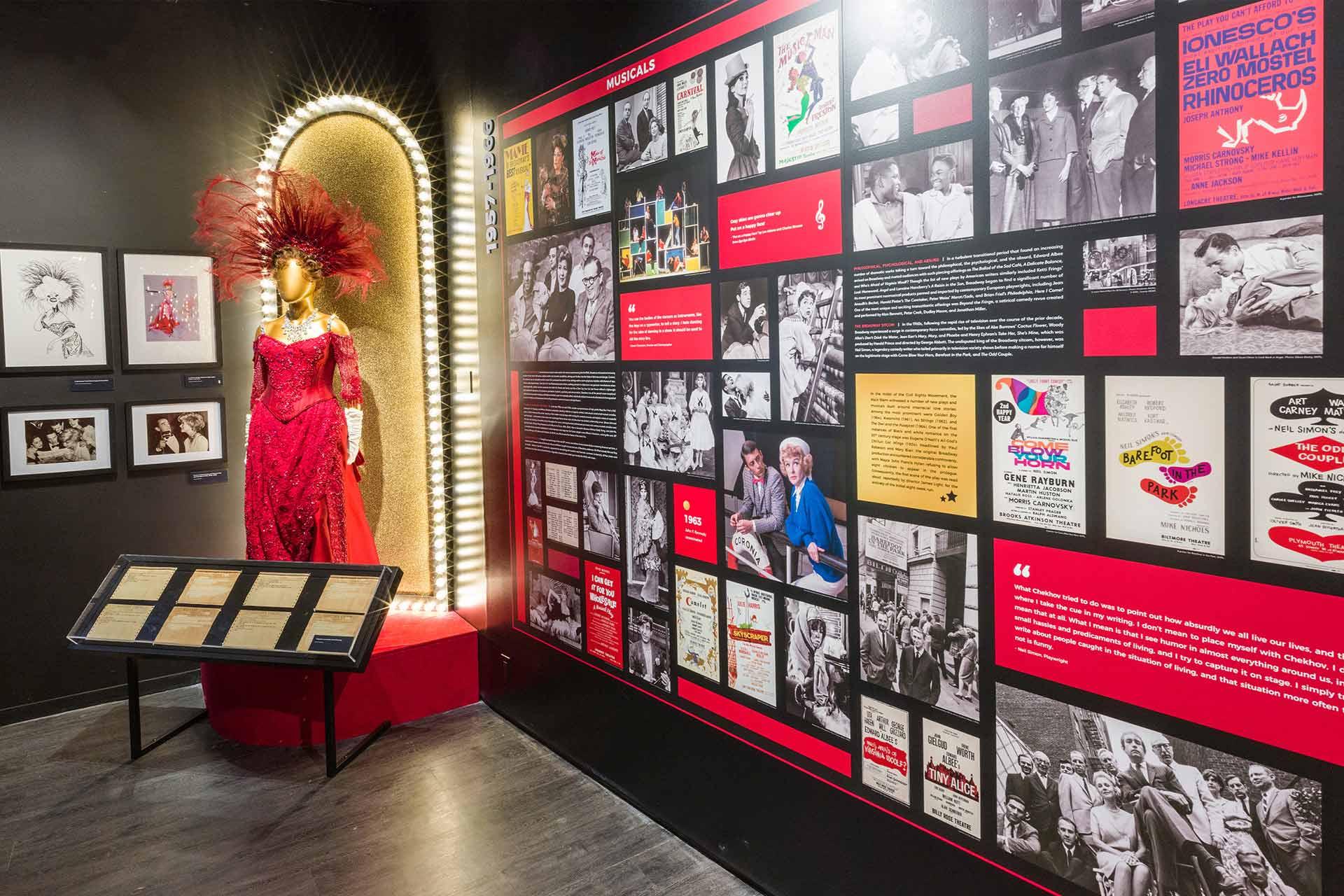 Museum exhibit featuring a sparkling red dress with a feathered headdress on a mannequin, spotlit next to framed photos, documents, and a large wall display of theater posters, images, and text about musicals.