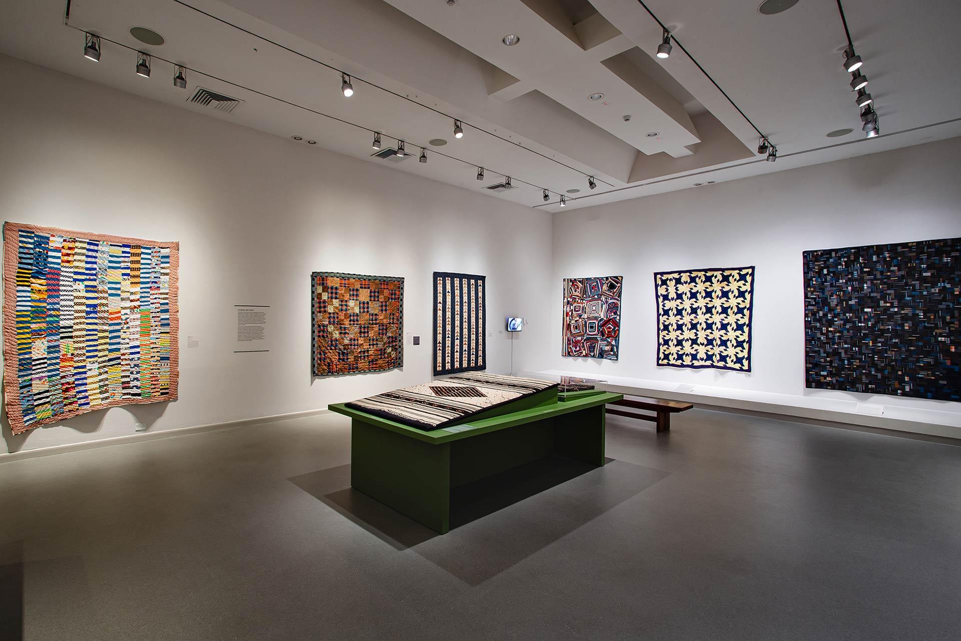A modern art gallery room displaying colorful quilts on white walls and on a central green table, with spotlights illuminating the textiles and a bench for visitors.