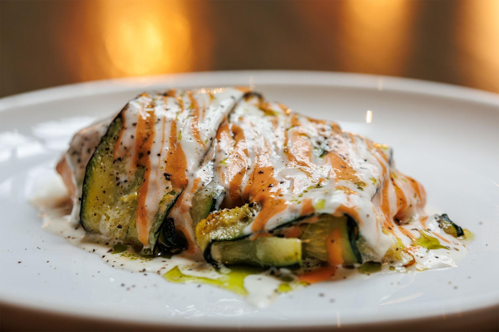 A close-up of a plated vegetable dish, featuring roasted zucchini slices drizzled with creamy white and orange sauces, garnished with ground black pepper and a touch of olive oil.