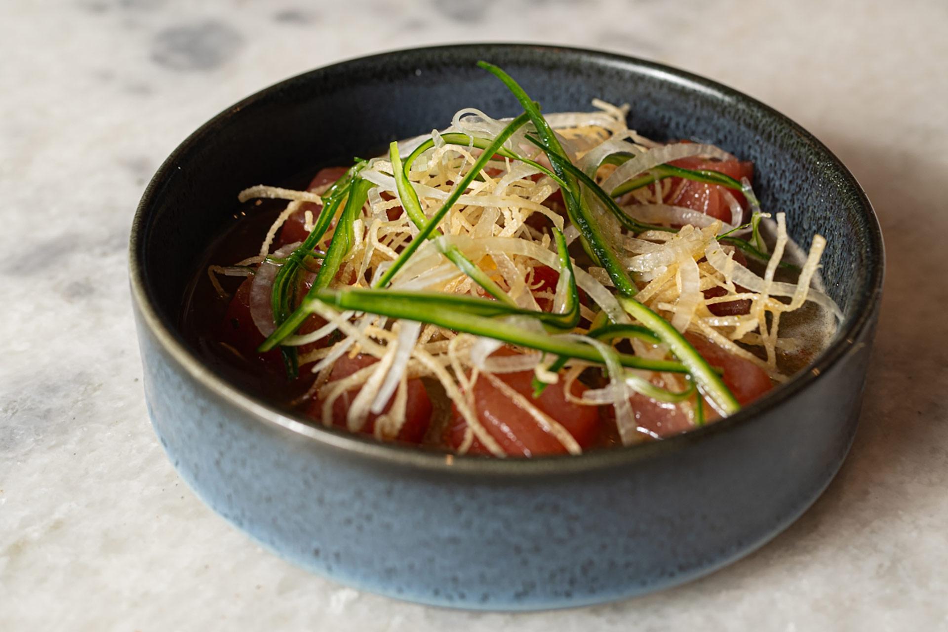 A blue bowl filled with thinly sliced raw fish, garnished with crispy fried strips, green herbs, and finely sliced onions, set on a light marble surface.
