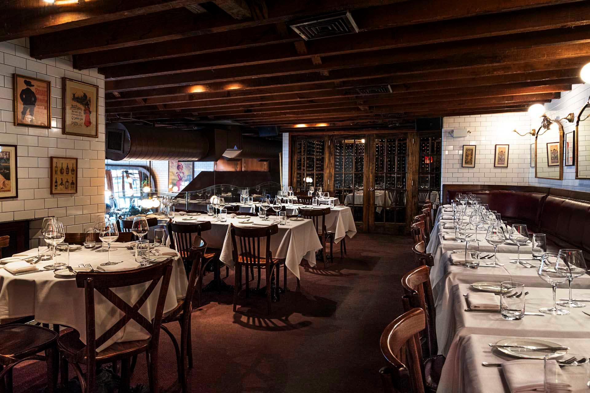 Dimly lit restaurant with wooden ceiling beams and white tiled walls. Several round tables are set with white tablecloths, glasses, and cutlery. Artworks adorn the walls, creating an intimate dining atmosphere.