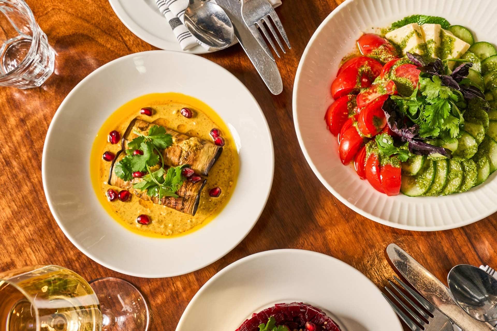 Overhead view of a wooden table set with plates of colorful dishes: a yellow curry with grilled eggplant and pomegranate seeds, a fresh salad with tomato, cucumber, and avocado, plus a glass of white wine and cutlery.